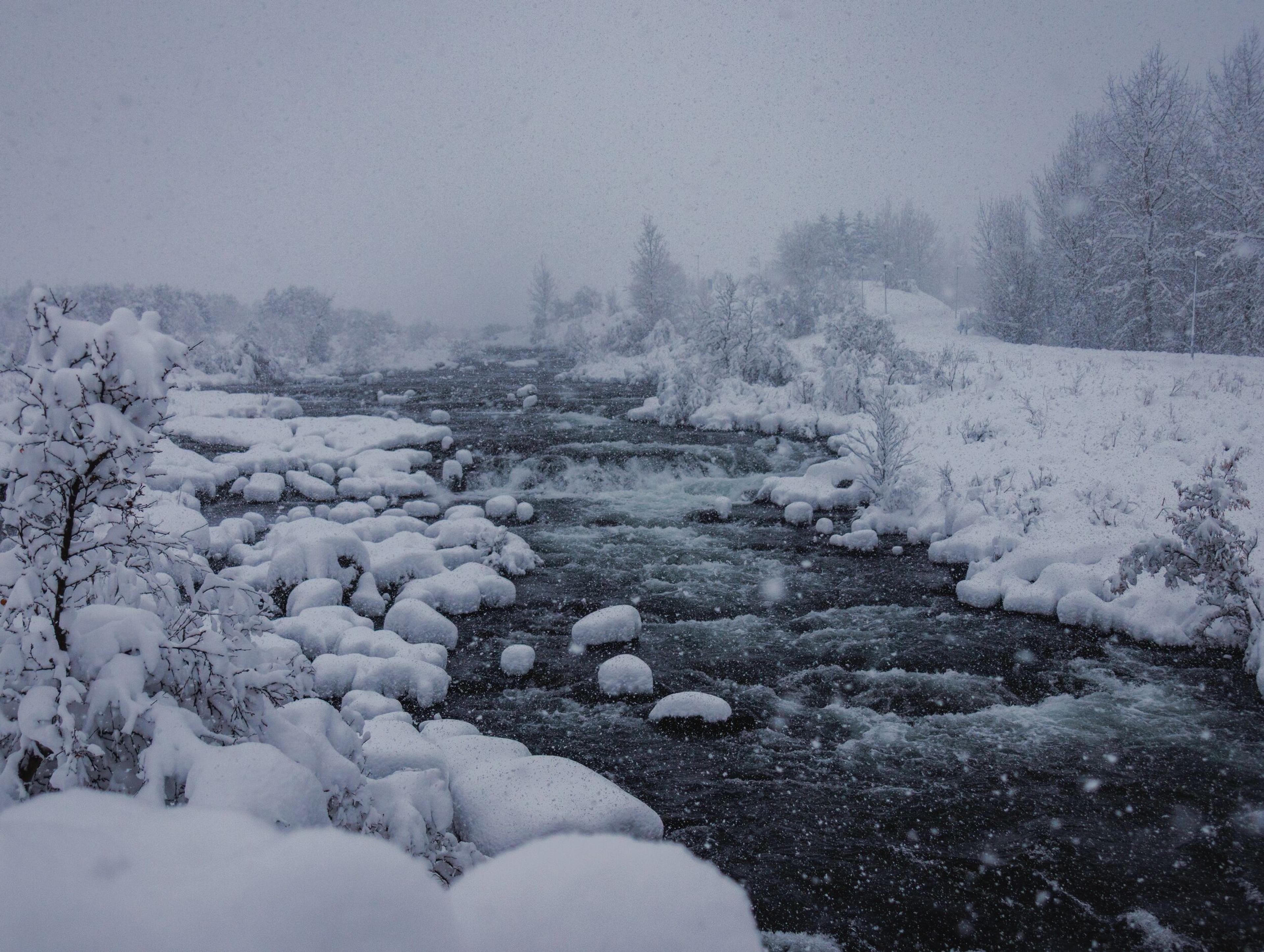A dark, turbulent river flows through a heavily snow-covered winter landscape during a snowfall.