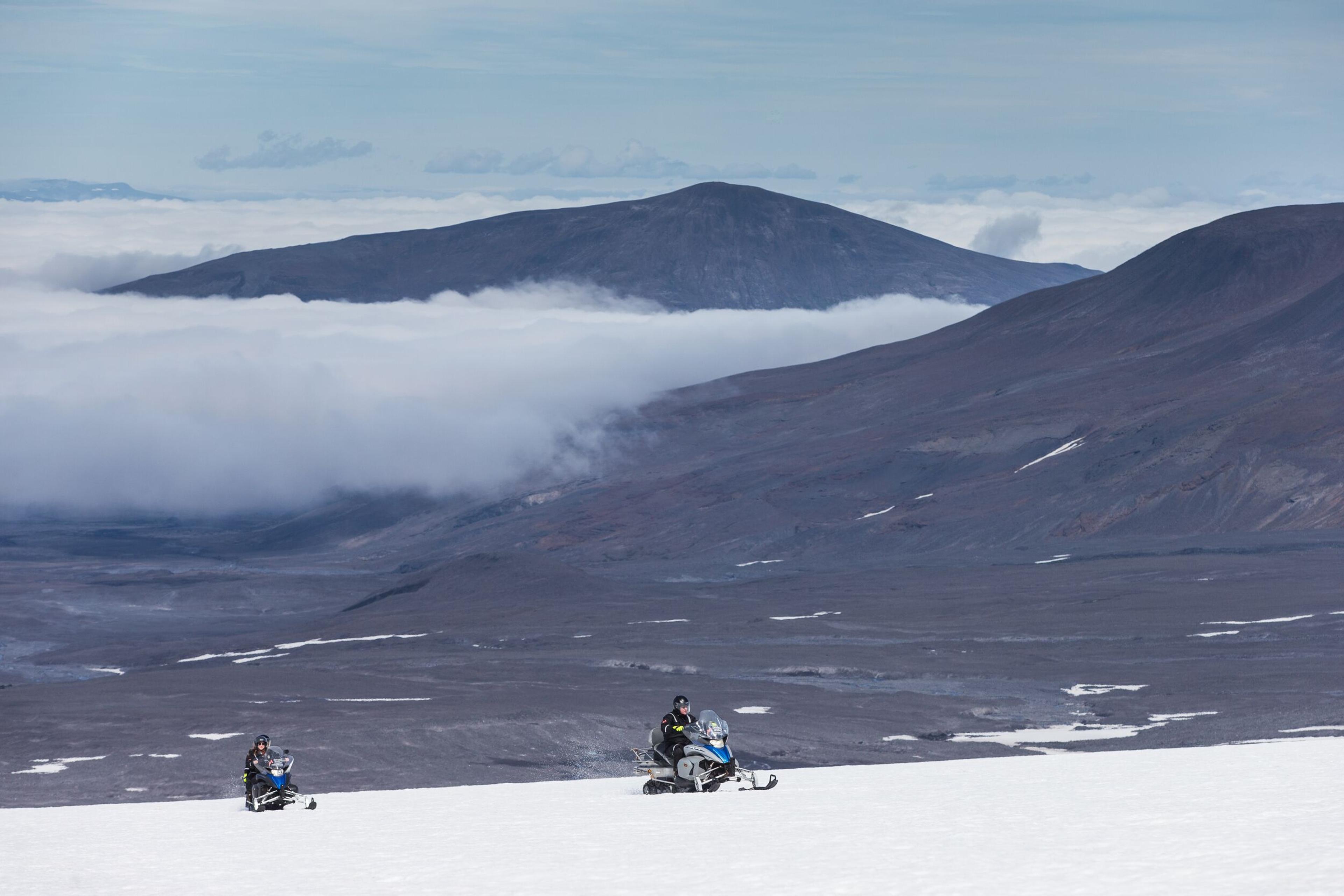 Two snowmobilers embarking on an adventure across the Langjökull glacier, with a rugged brown mountain and dramatic clouds in the background