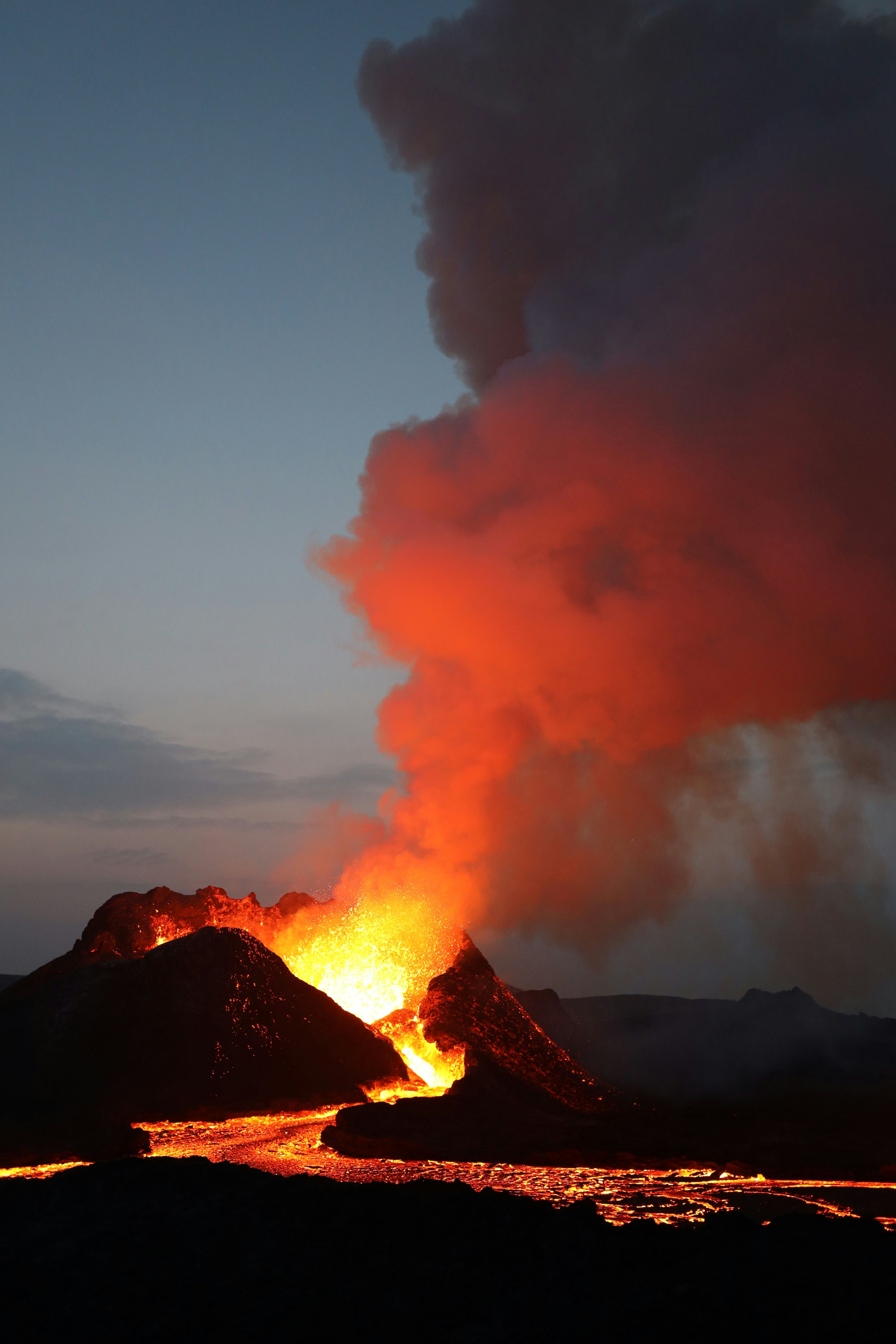 Fiery volcanic eruption on the Reykjanes Peninsula near Grindavik, with molten lava flowing down the dark landscape and thick smoke billowing into the twilight sky.