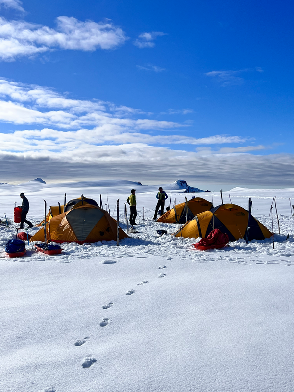 Explorers camping on Vatnajökull ice cap, with bright yellow expedition tents and a few people in cold-weather gear. The footprints in the snow lead towards the tents, and the sky is clear with some scattered clouds, creating a striking contrast between the white snow and the deep blue sky