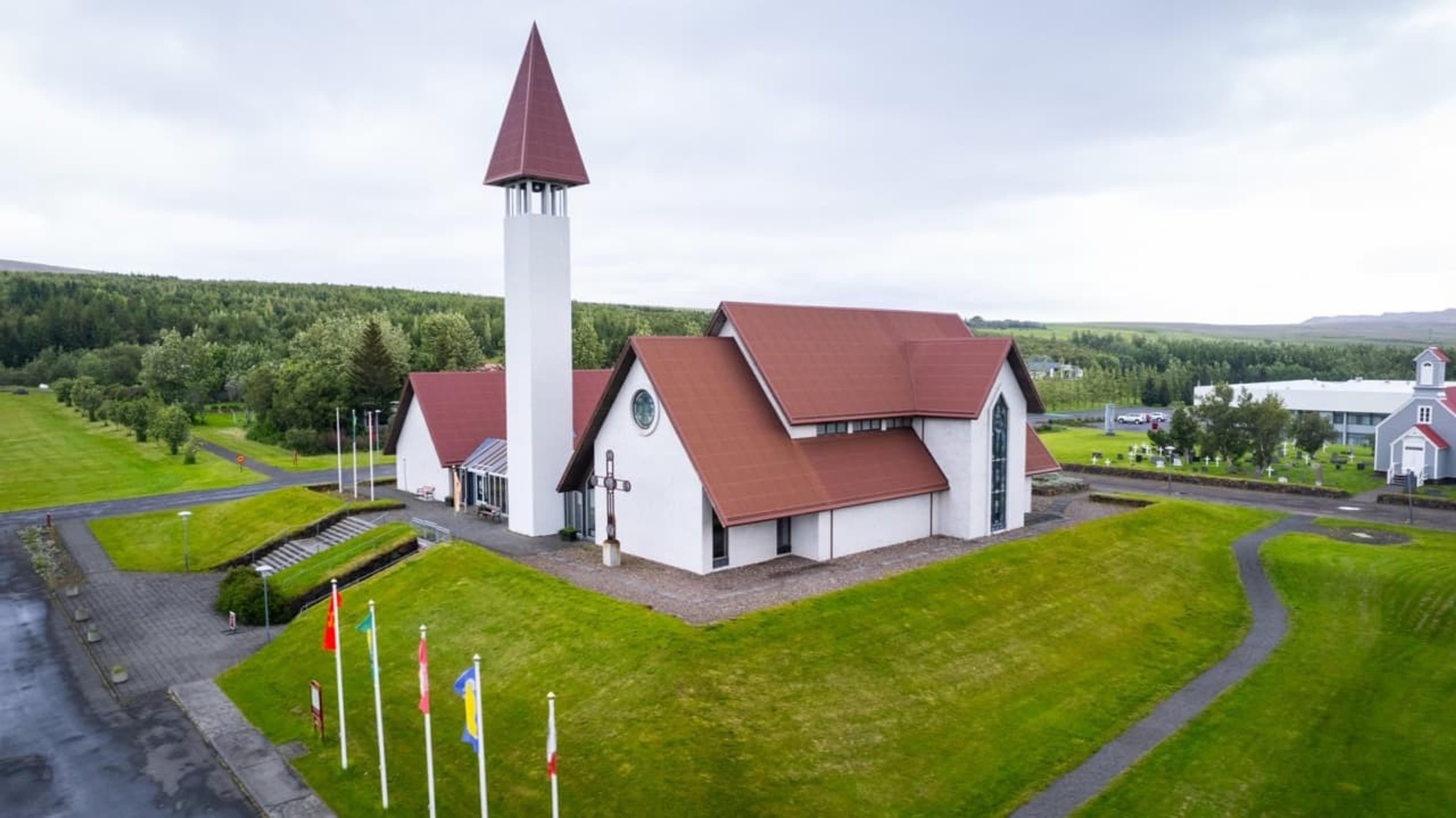 A modern white church with a red roof and tall bell tower, surrounded by manicured green lawns and trees, located in Reykholt, Iceland.