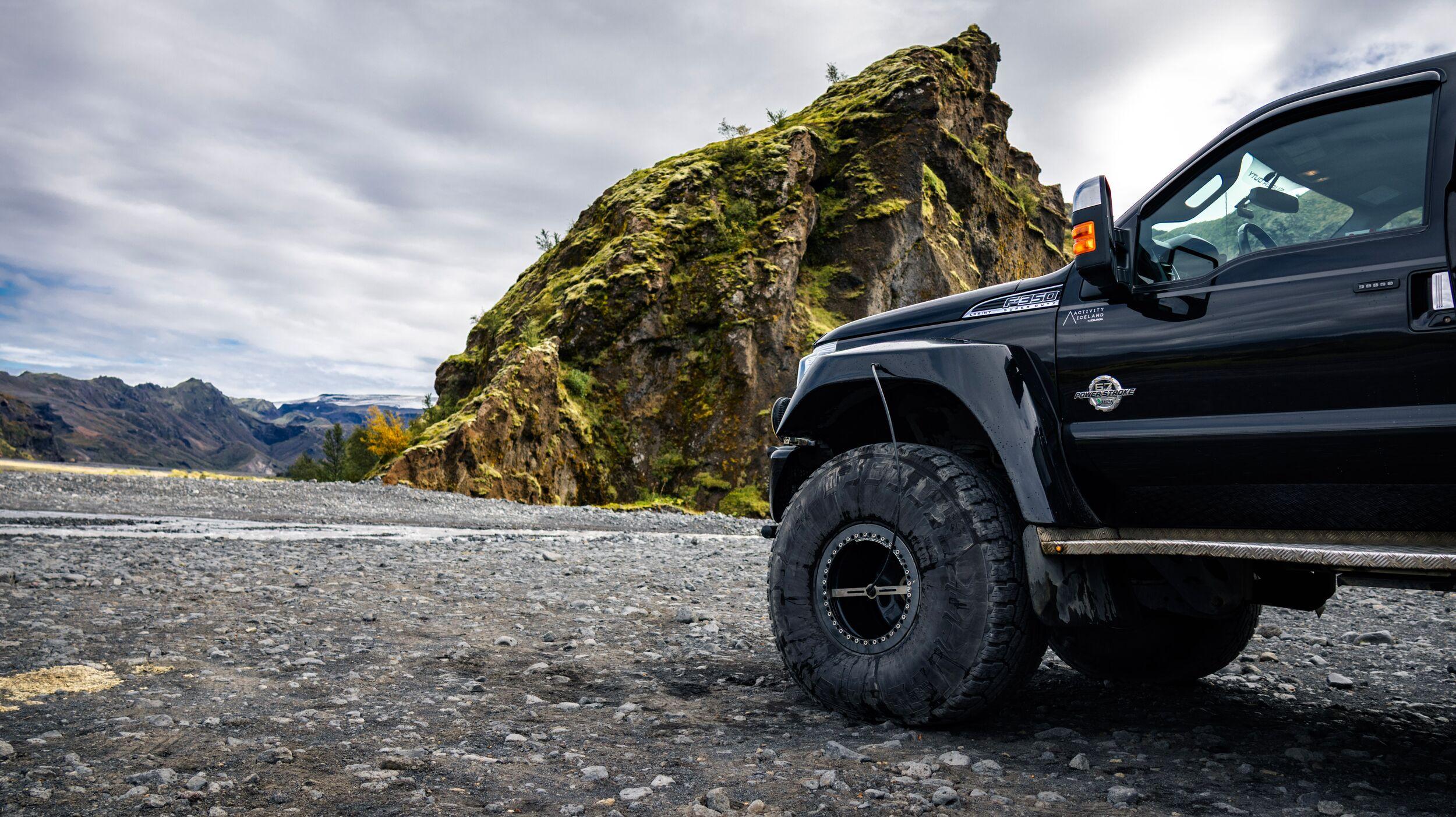 Black off-road pickup truck with massive tires on a rocky path, with a moss-covered mountain and distant peaks under a cloudy sky.