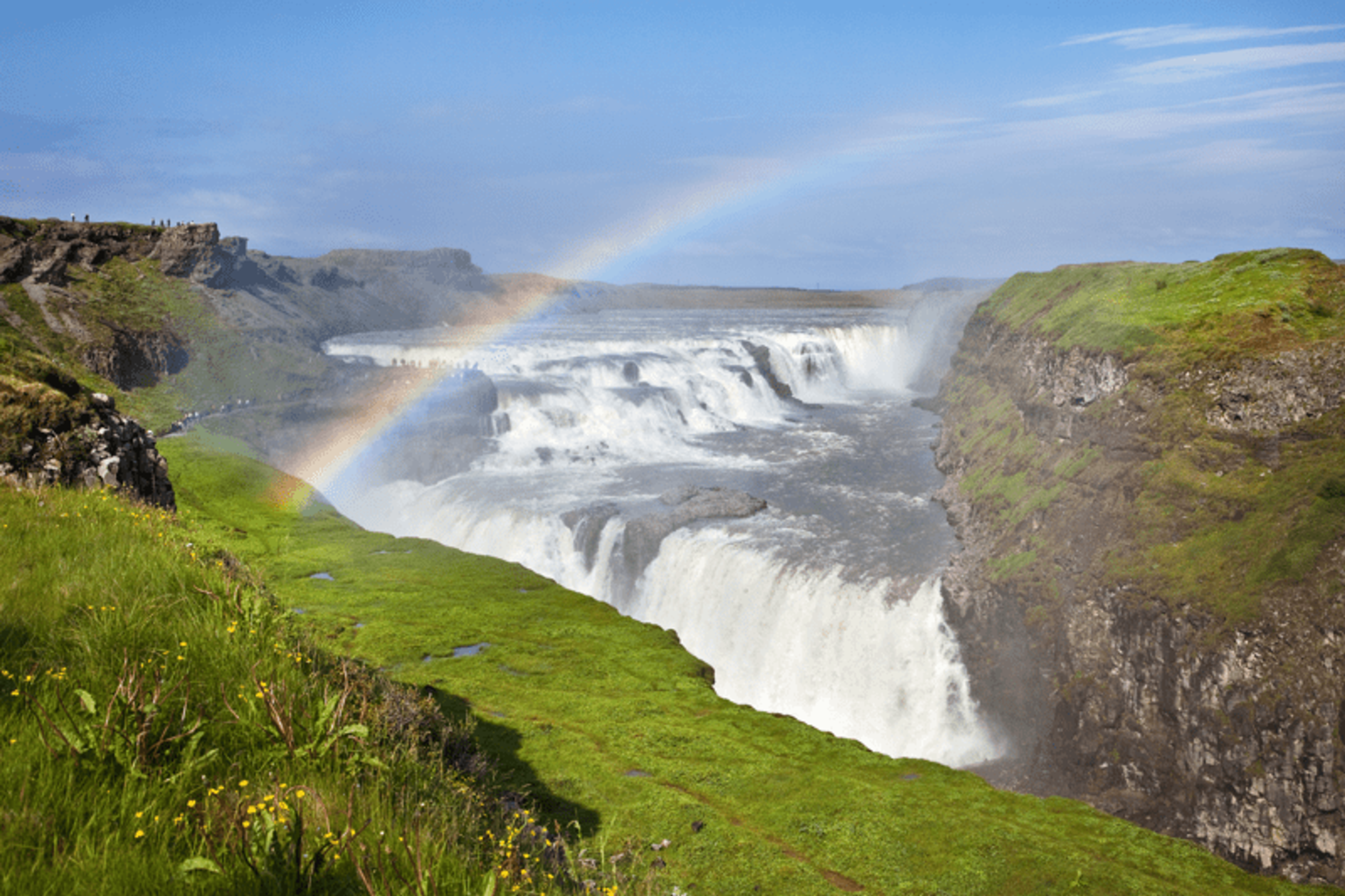 Rainbow over Gullfoss waterfall cascading between green cliffs.