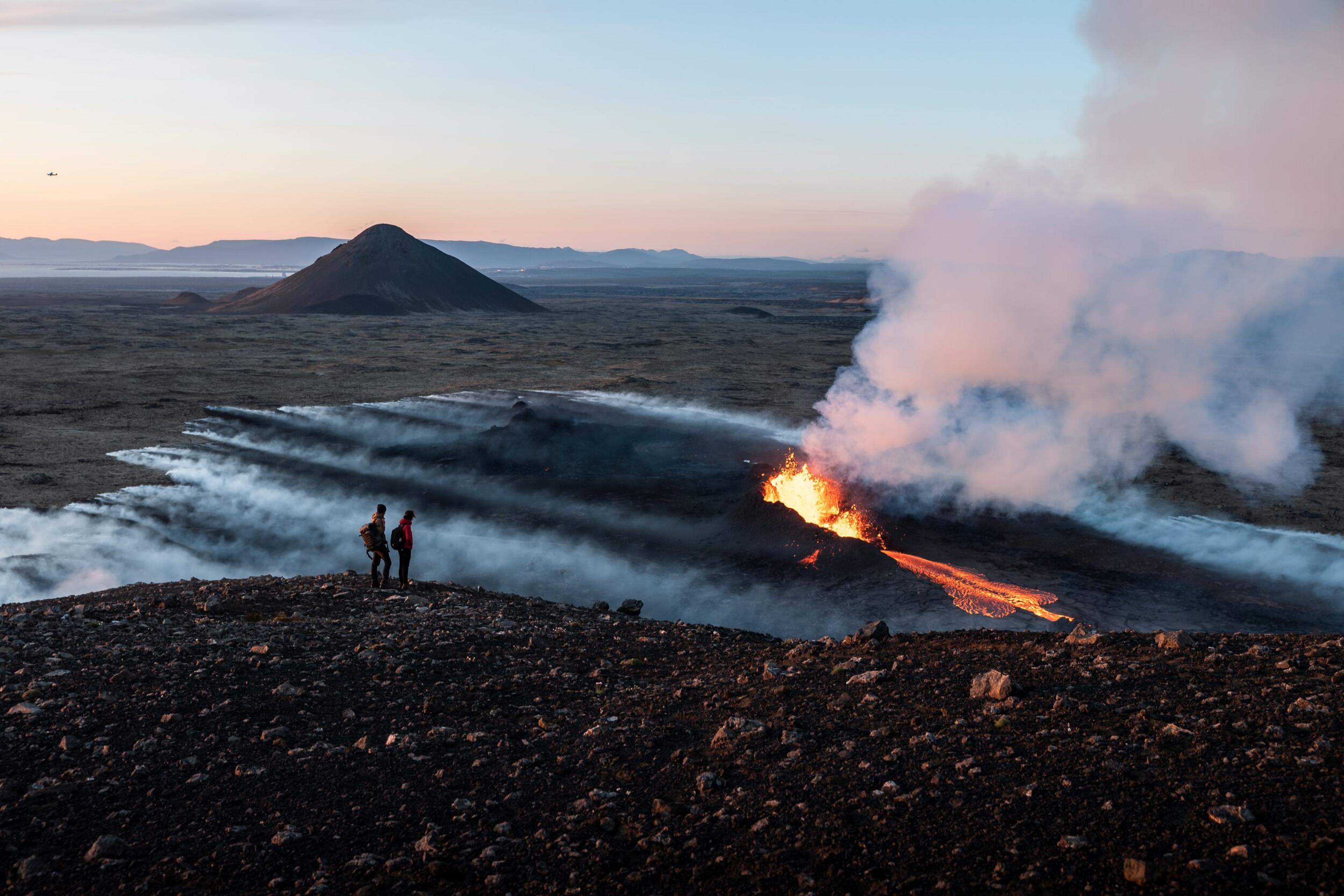 Two people stand on a ridge, watching lava flow from a volcanic fissure, with smoke rising and a distant mountain under a twilight sky.