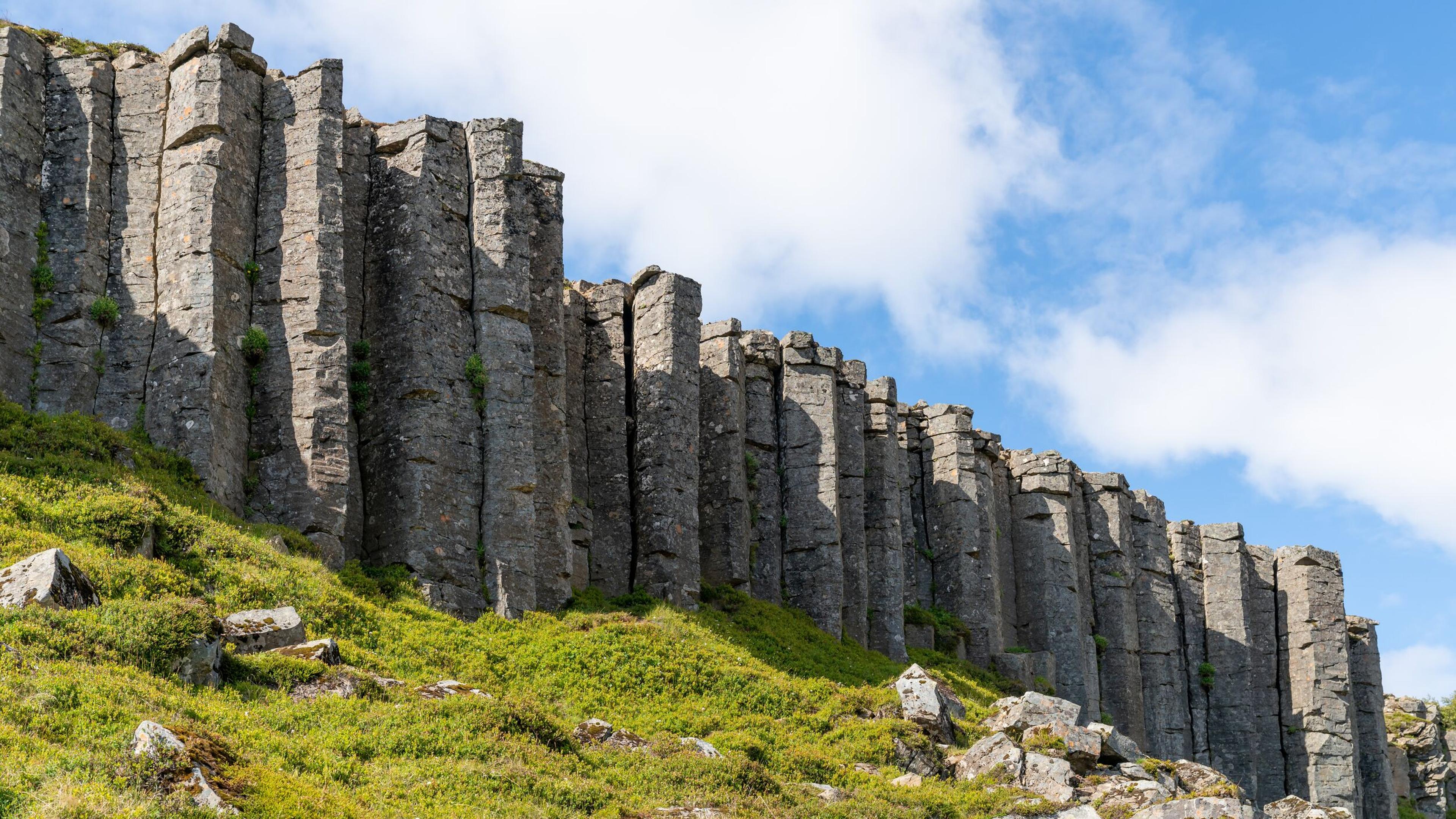 A striking view of Gerðuberg basalt columns in Iceland, showcasing tall, perfectly aligned hexagonal rock formations rising from a lush green hillside under a bright blue sky.