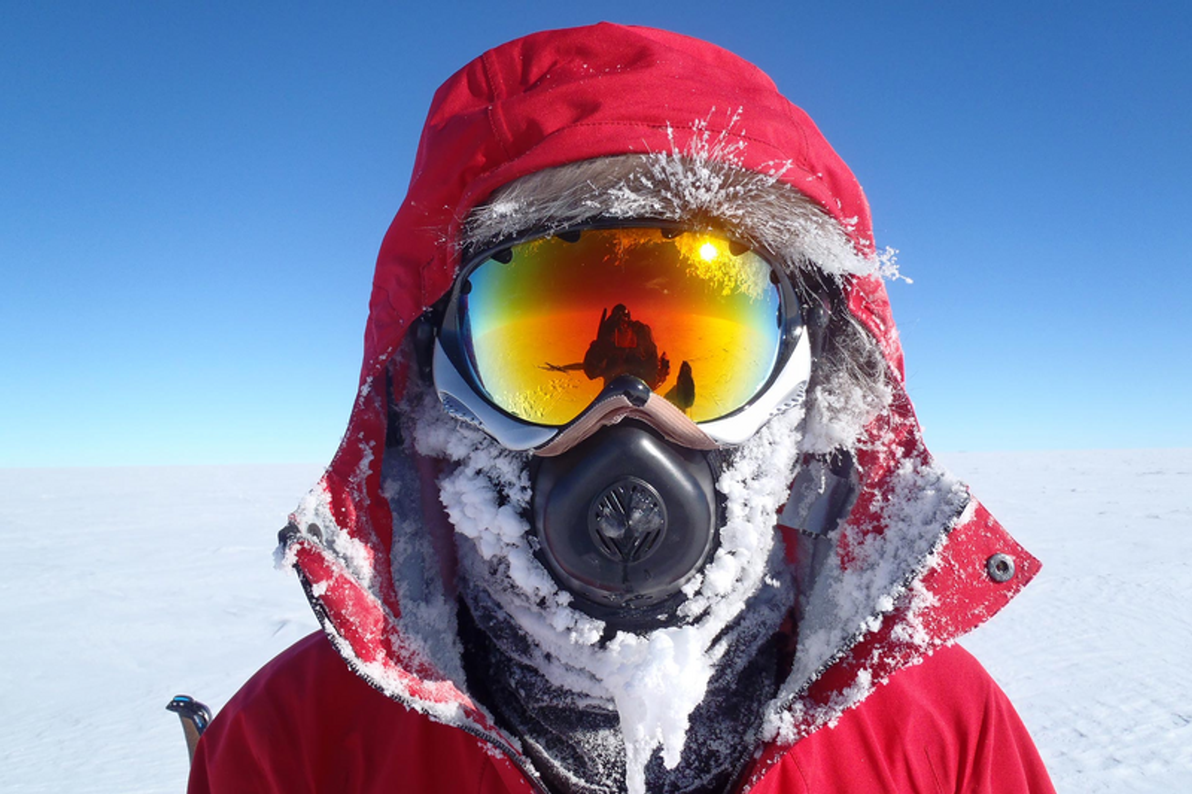 Person wearing a red hooded parka and a full-face ski mask, covered in snow and ice, braving the cold elements.