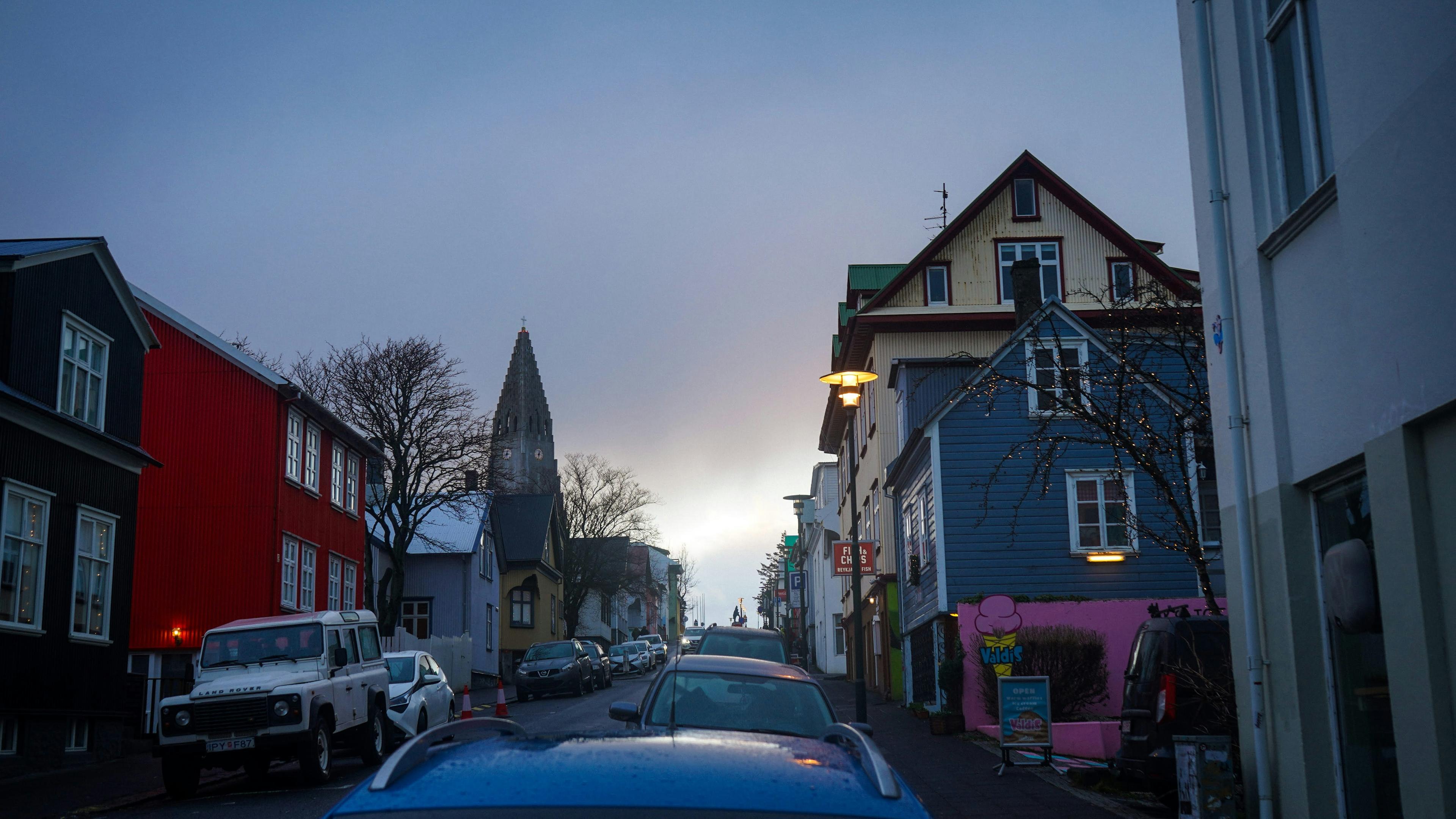 Street view in Reykjavik with colorful houses, parked cars, and Hallgrímskirkja church visible in the distance at dusk.