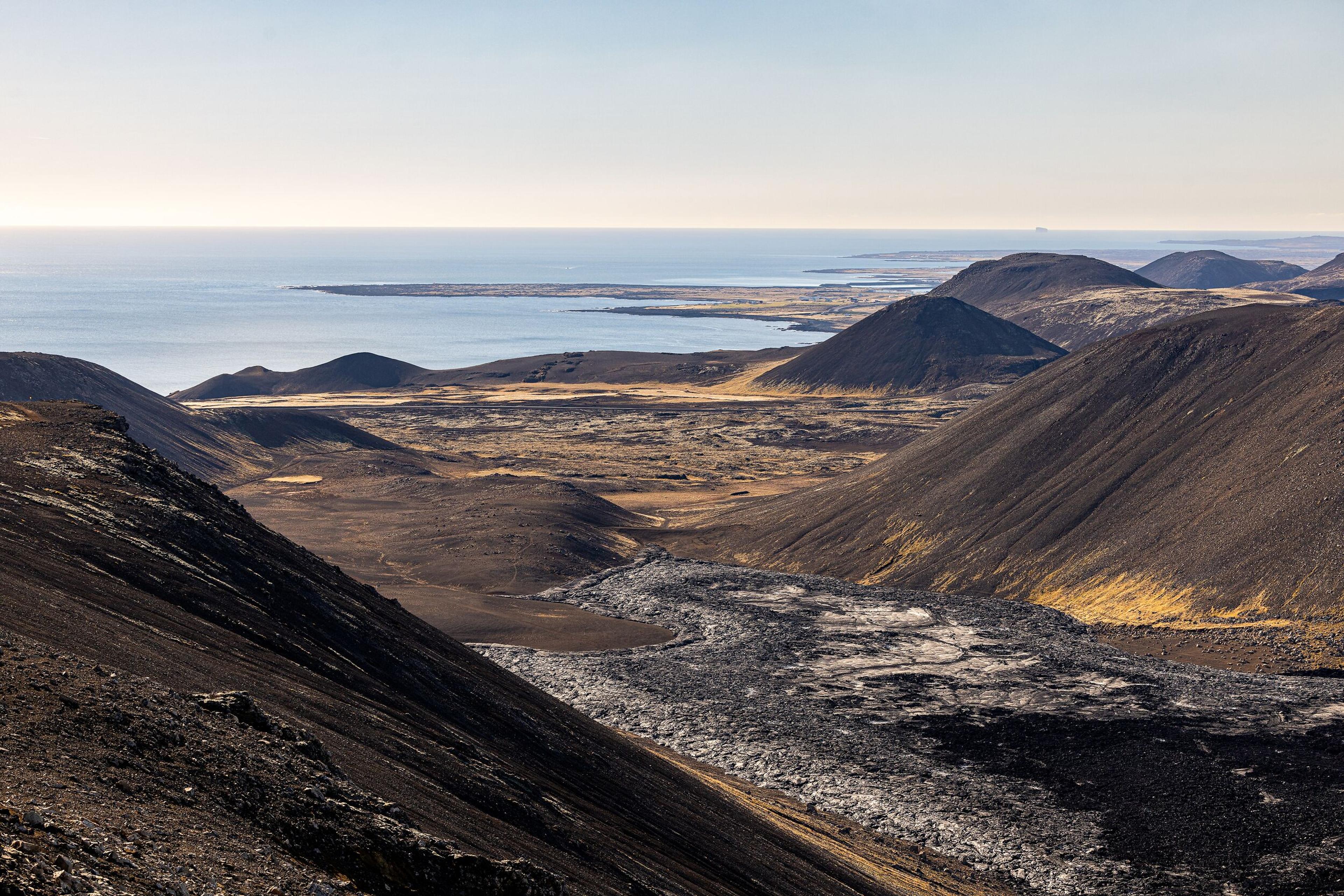 A sweeping view of a volcanic landscape with dark lava fields, rolling hills, and the ocean stretching into the horizon under a clear sky.