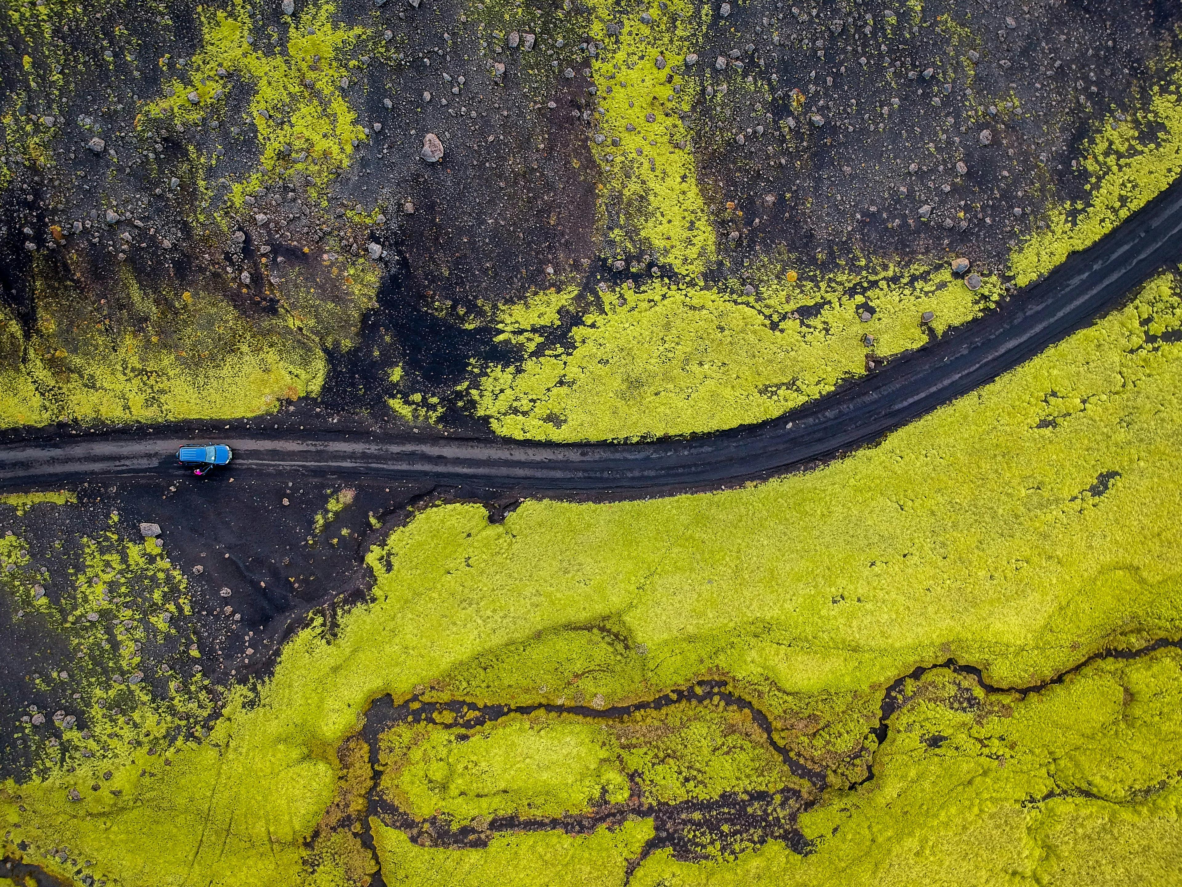 Aerial view of a blue car on a rugged road cutting through vibrant green moss-covered landscape in Iceland.