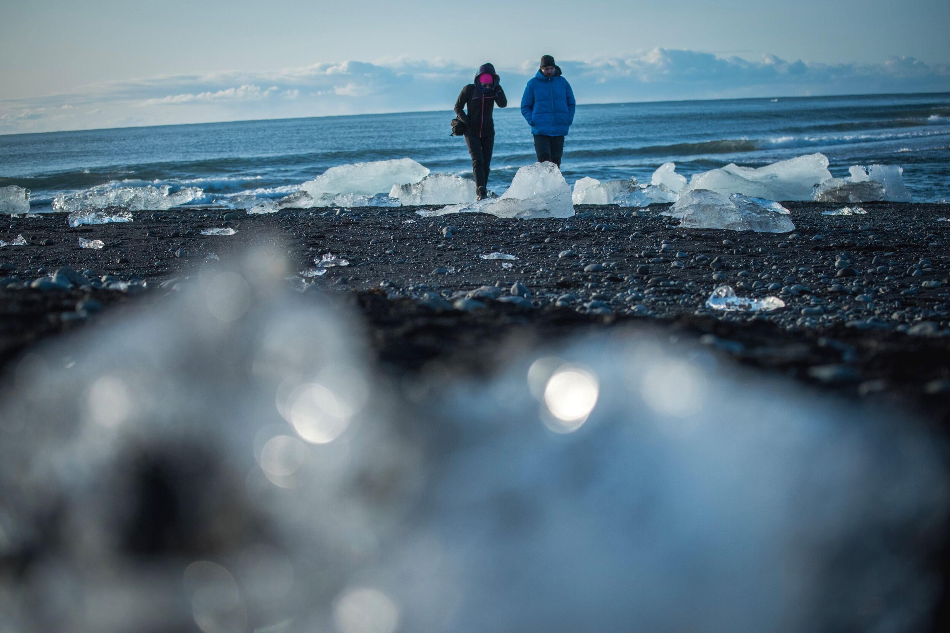 Two people walk on a black sand beach covered in large ice chunks, with ocean waves in the background.