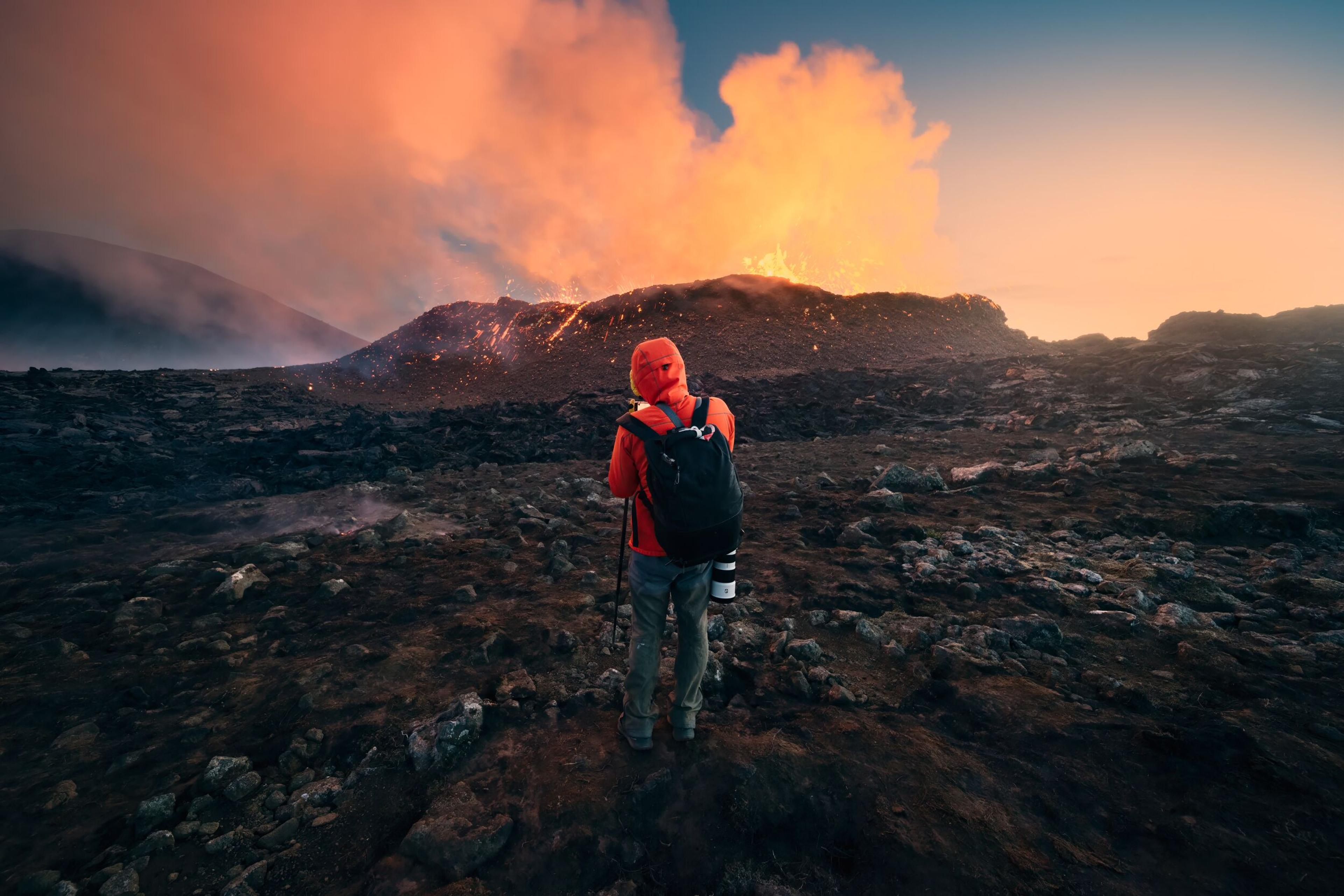 A person in a red jacket stands at a safe distance, witnessing the raw and mesmerizing power of an active volcanic eruption in Iceland, with the twilight sky glowing from the lava's reflection