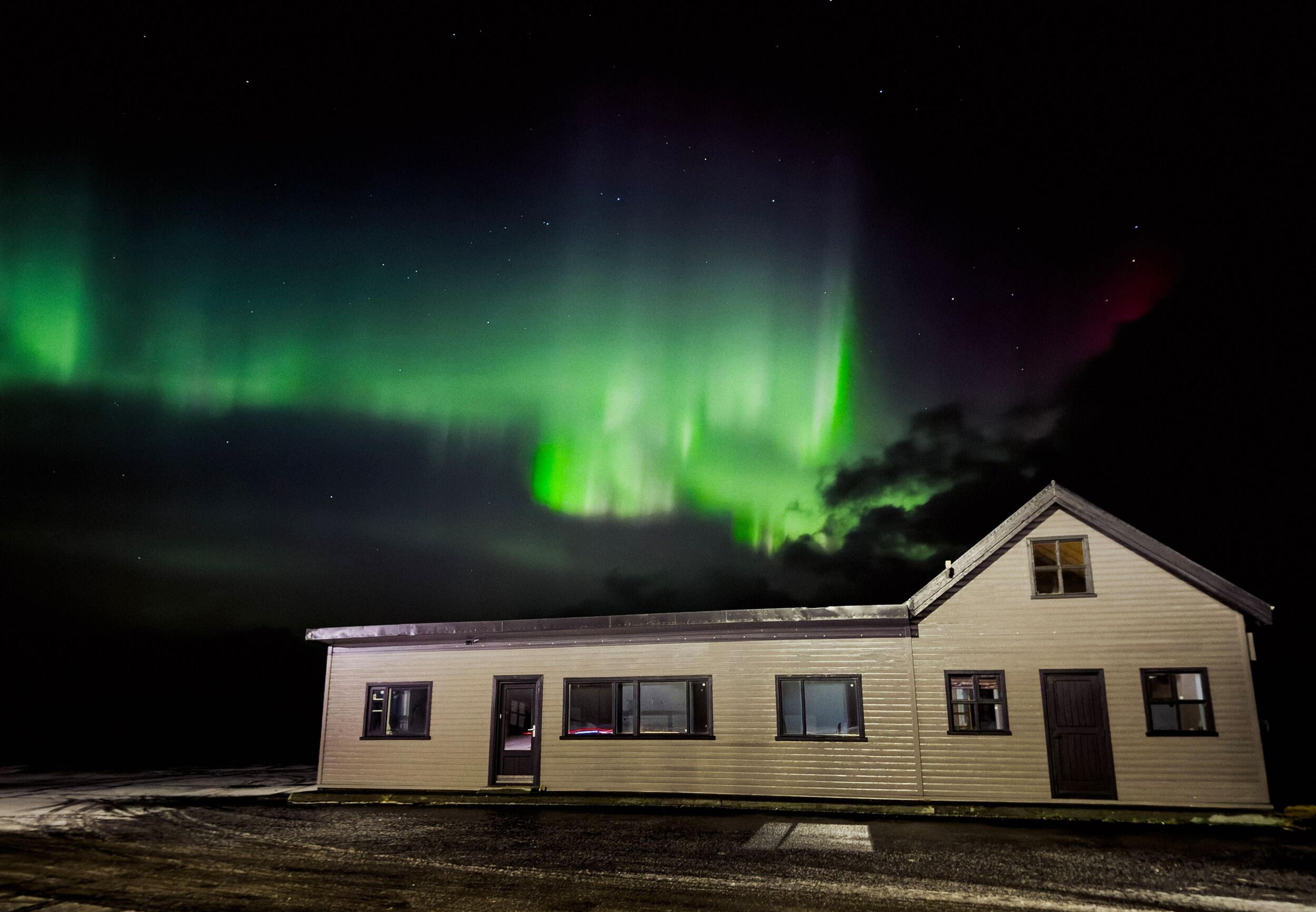 A house with illuminated windows beneath a vibrant green aurora borealis.