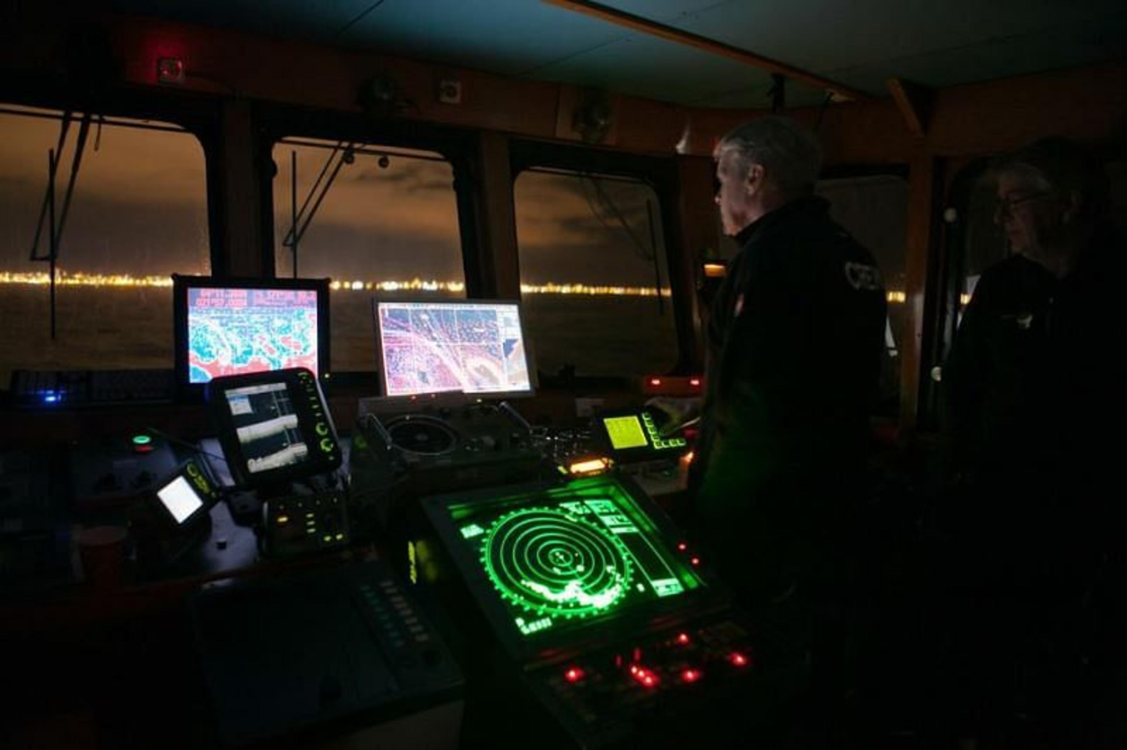 Ship's bridge at night with two crew members, glowing navigation screens, and distant city lights.