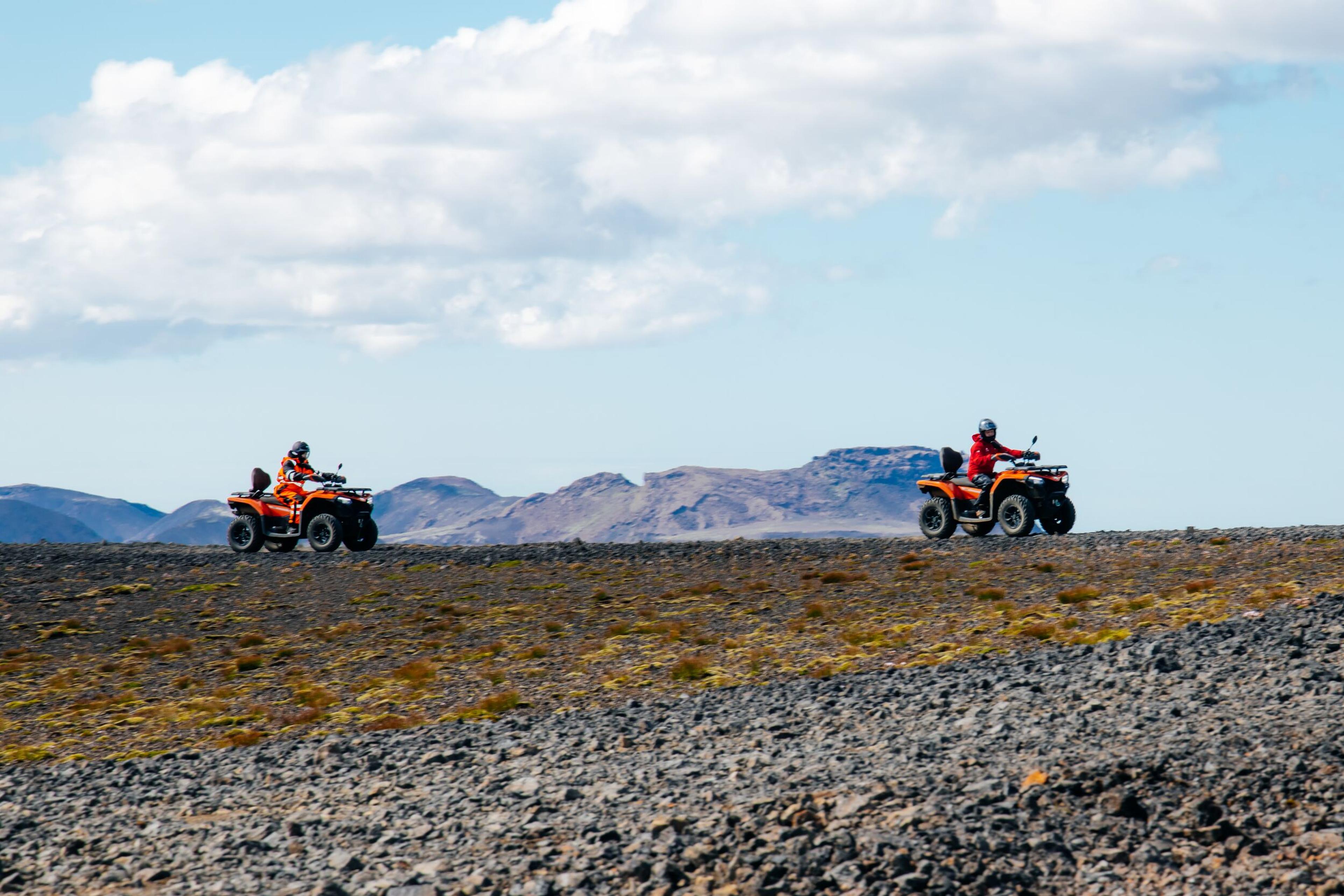 Two people riding orange ATVs across a rocky, barren landscape.