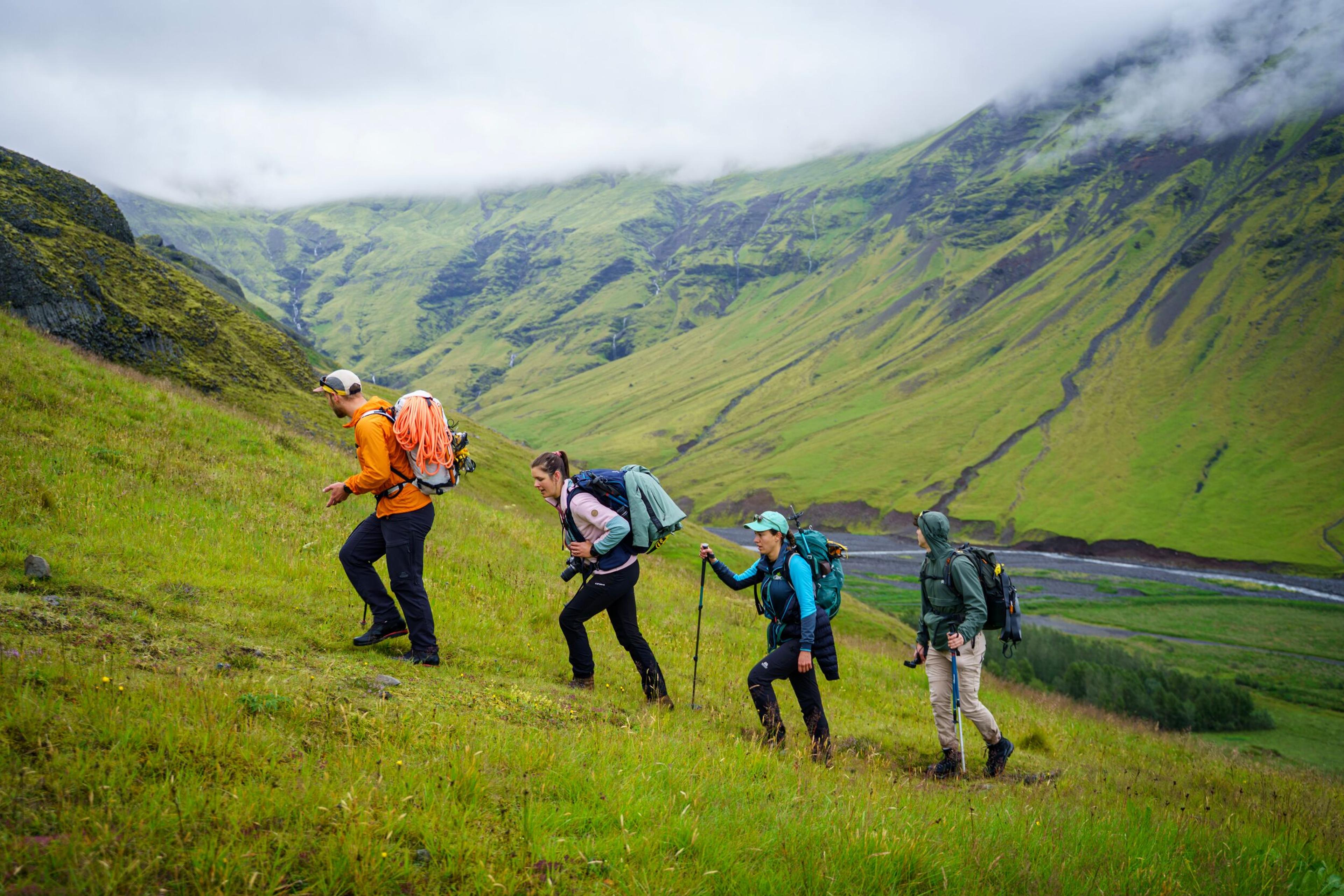 Four hikers ascend a lush green hillside with mountains and a river in the distance.
