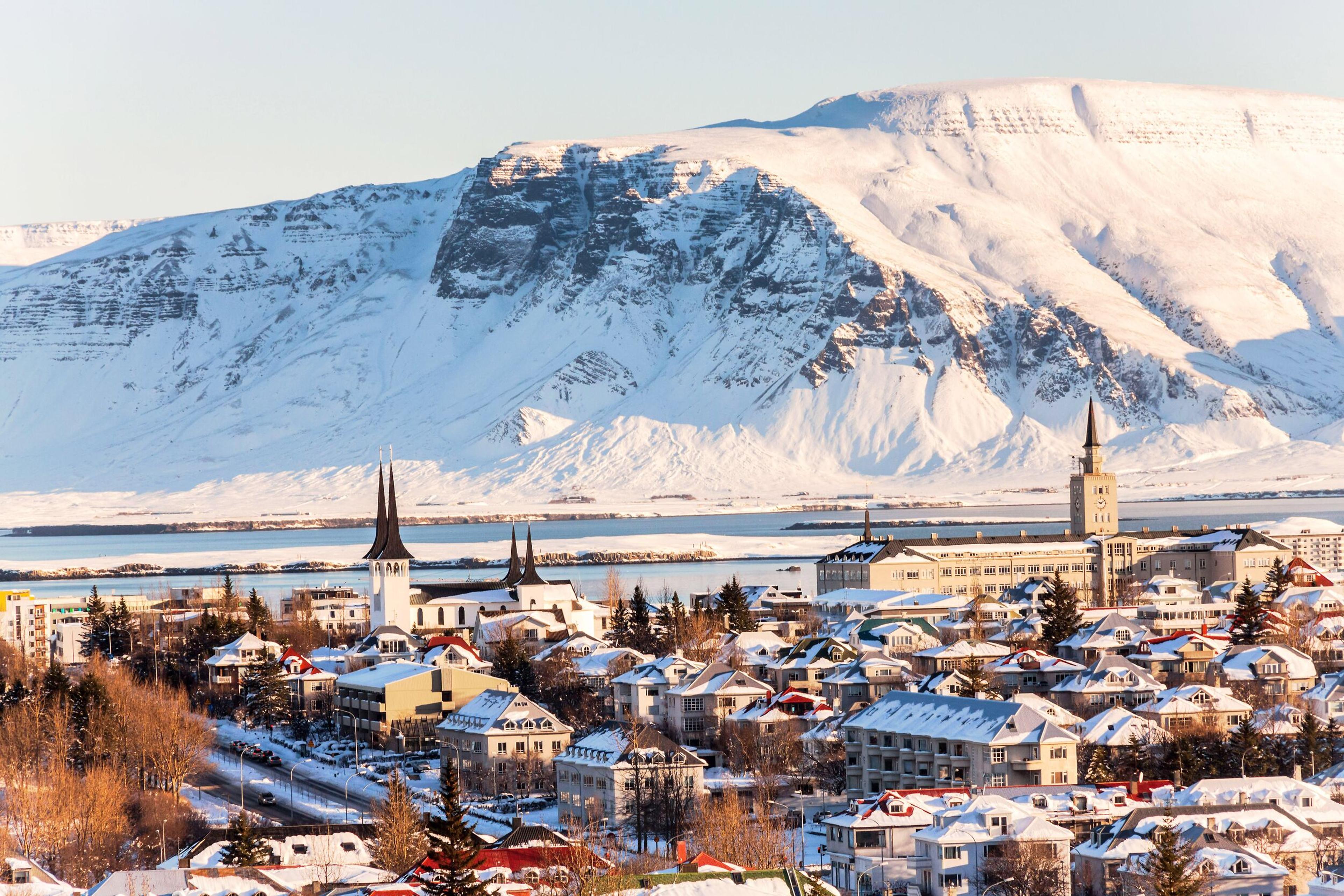 The snowy Reykjavík with distinctive architecture, including a tall church spire, against a backdrop of a mountain bathed in the warm glow of the sun.