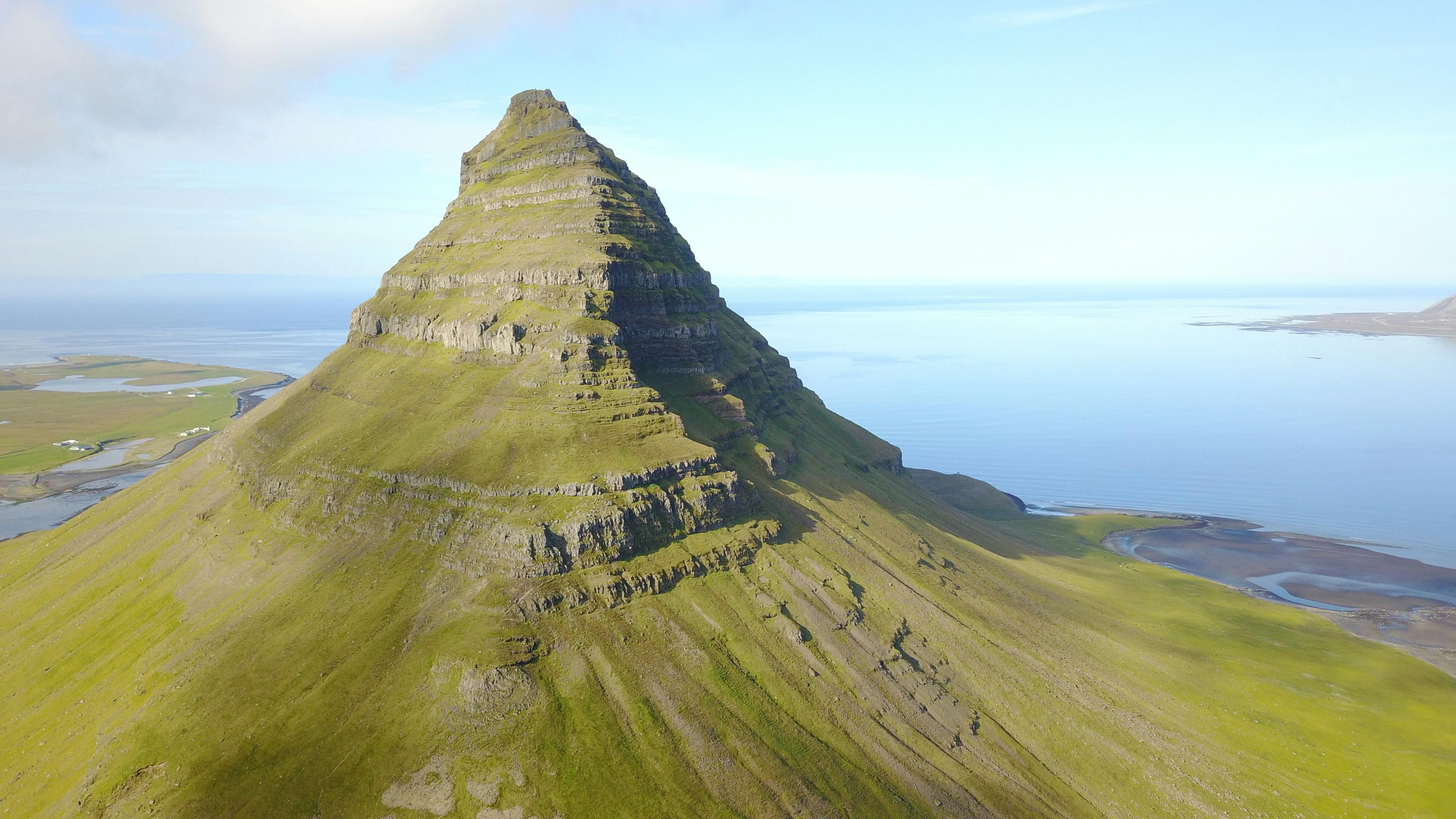 A stunning aerial view of Kirkjufell, Iceland's iconic pyramid-shaped mountain, covered in lush green grass and surrounded by calm coastal waters under a clear blue sky.