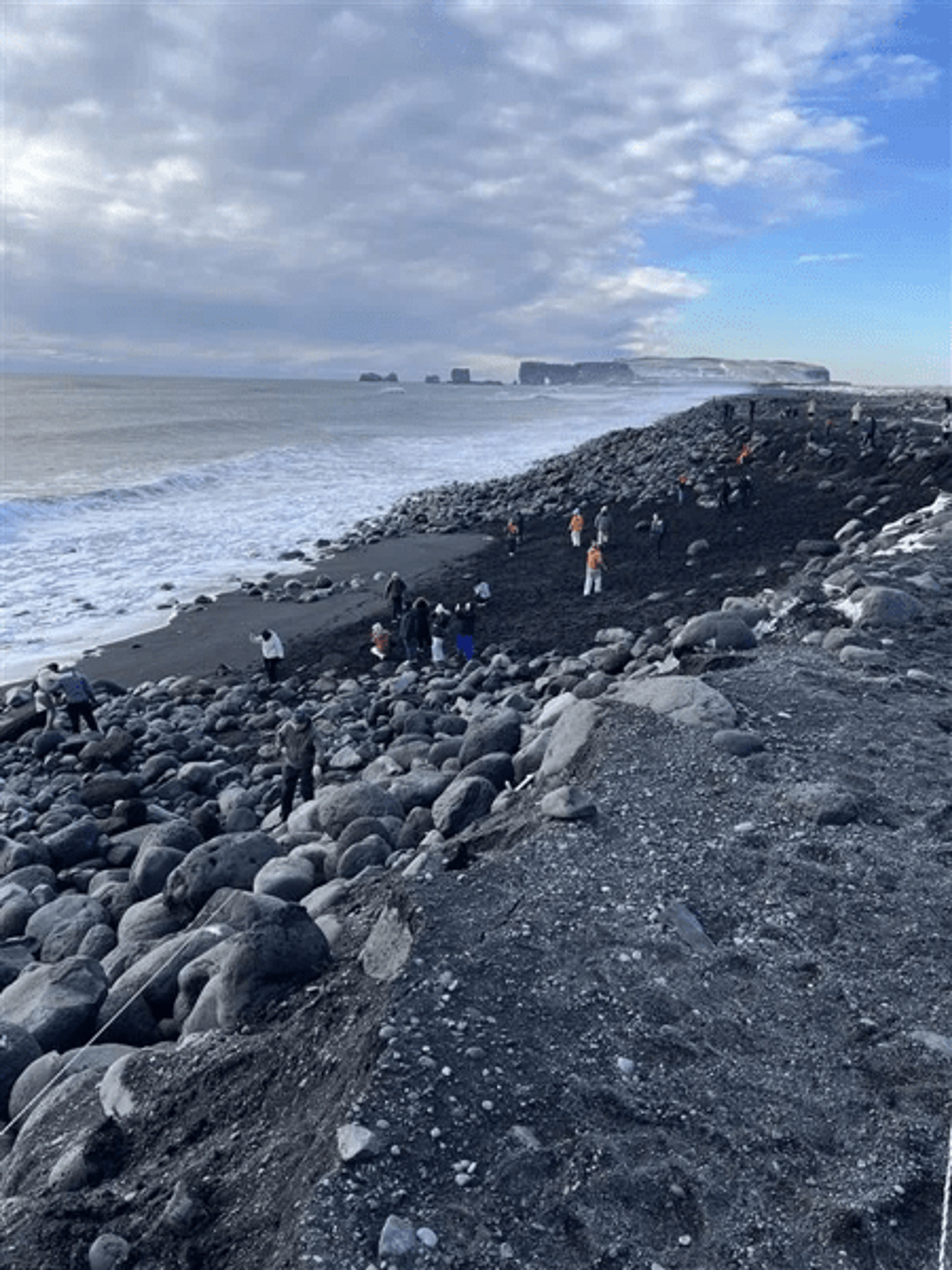 Group of people on a black sand and rock beach with ocean waves and distant cliffs under a cloudy sky.