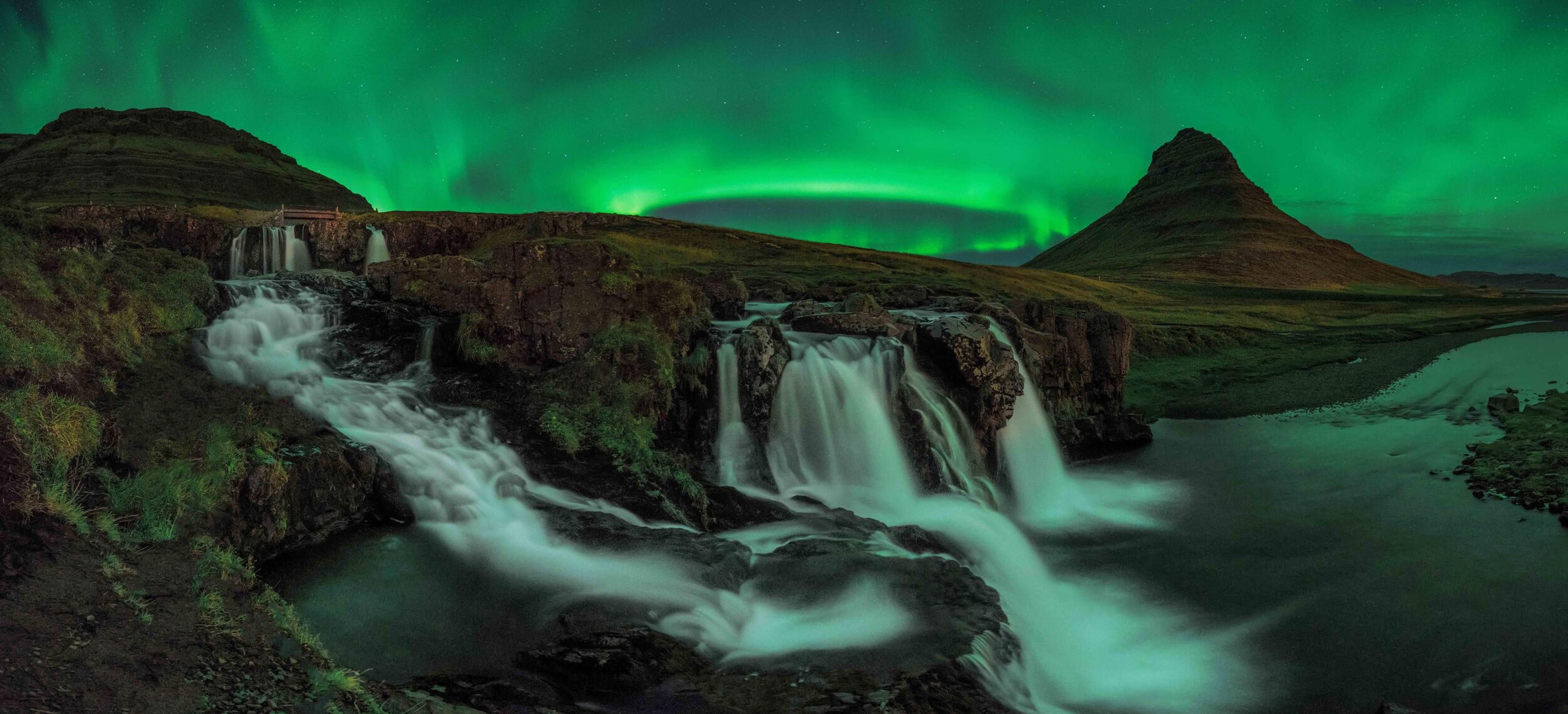 Green aurora borealis illuminates Kirkjufell mountain and a multi-tiered waterfall.