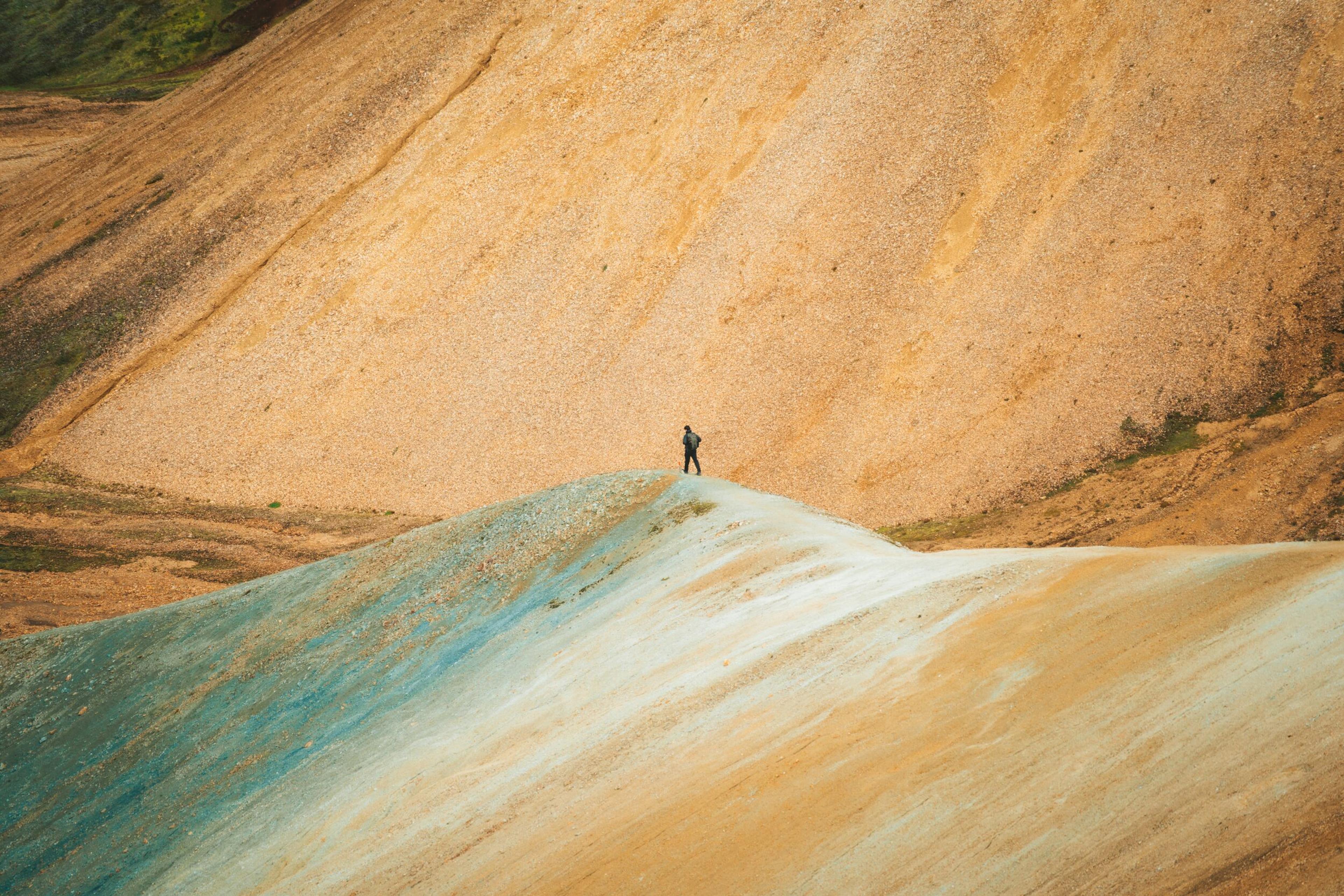 A hiker stands on a colorful mountain ridge with sandy and blue-green slopes.