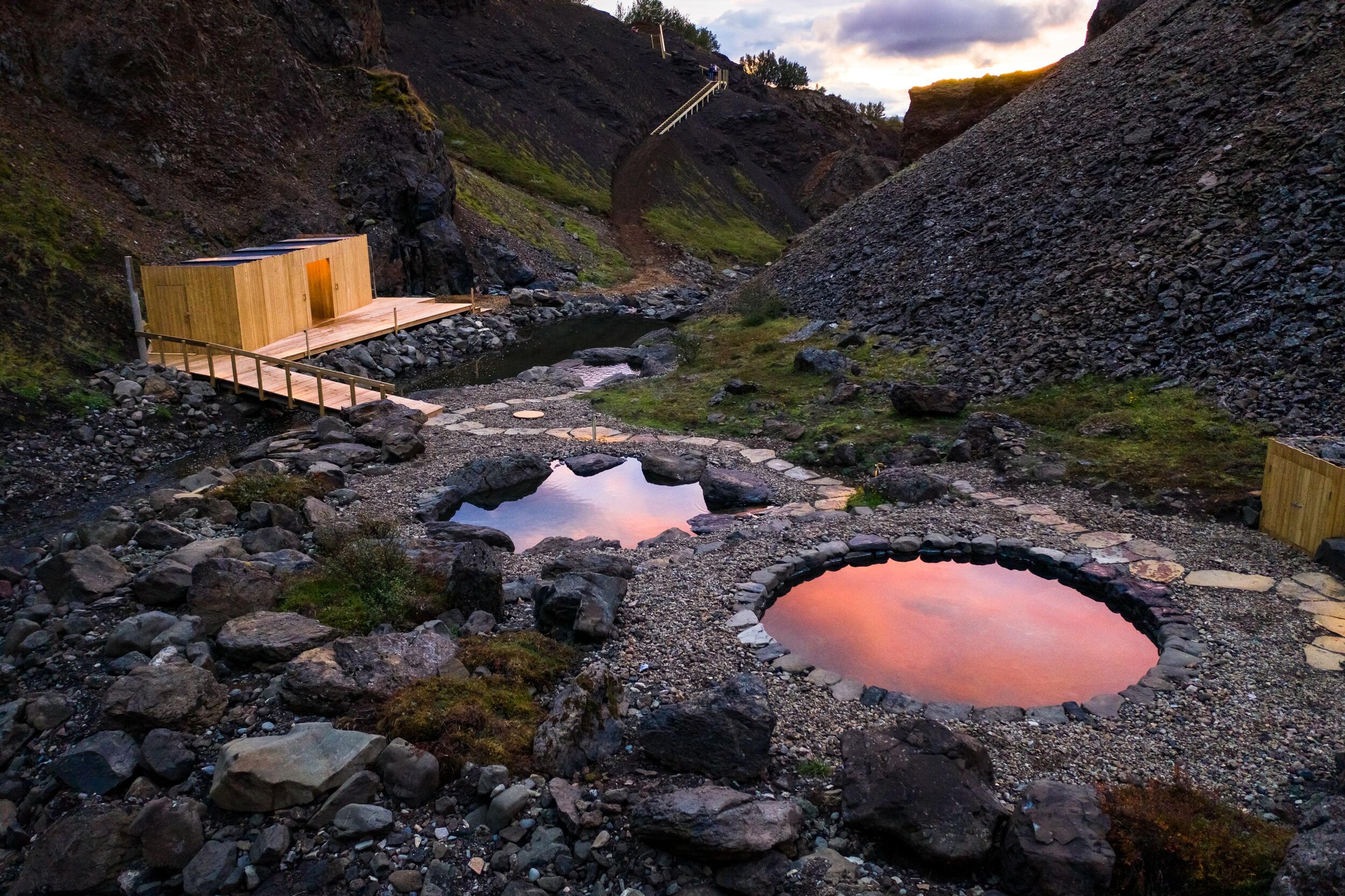 Natural hot springs at Húsafell on the Silver Circle, surrounded by rocky terrain and greenery. The pools reflect the pink and orange hues of the sky at sunset, with a small wooden structure and walkway nearby, blending into the serene landscape.