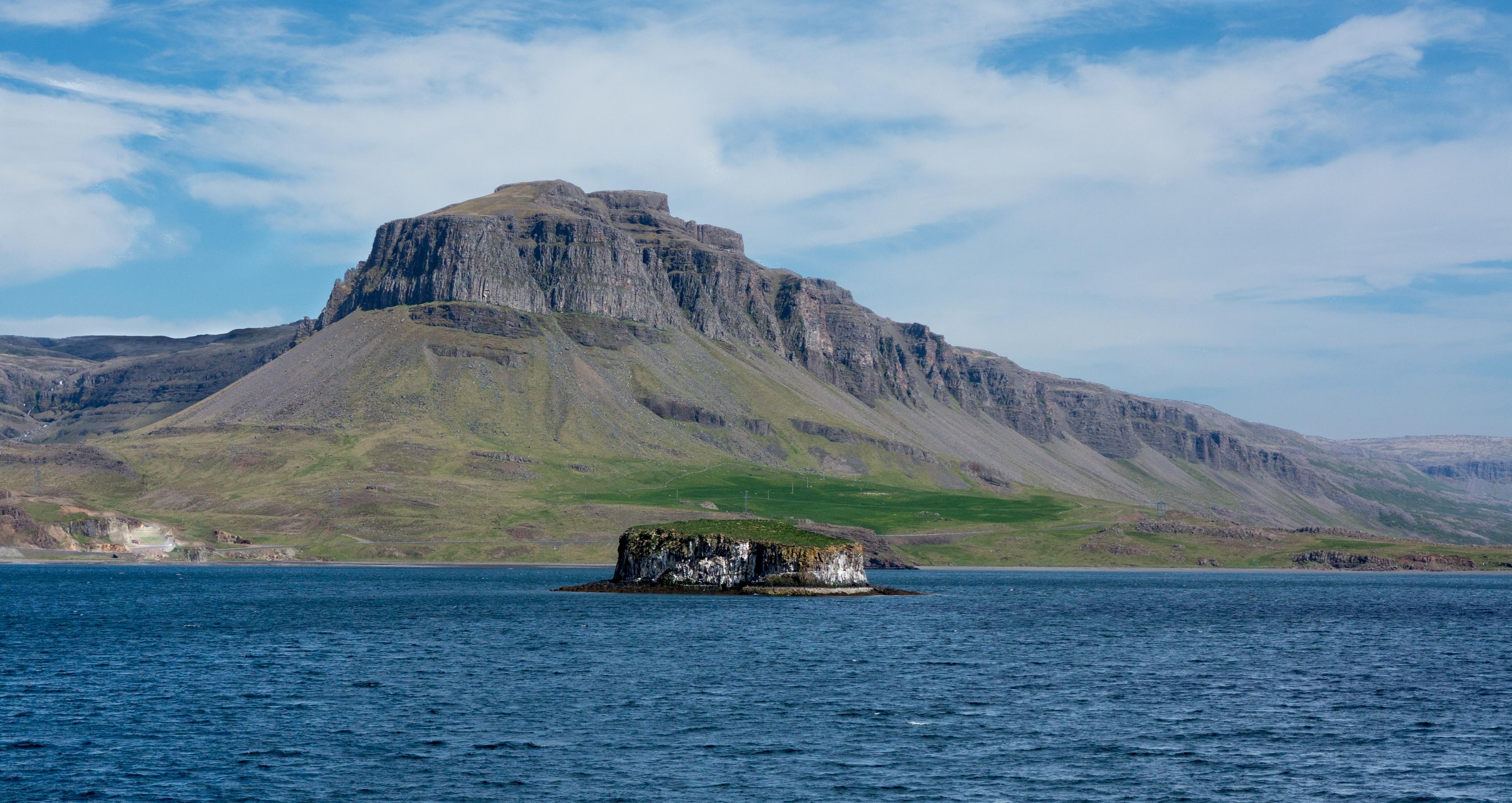 A scenic mountain towers over the calm waters of a fjord, its slopes covered with lush greenery against a clear sky.
