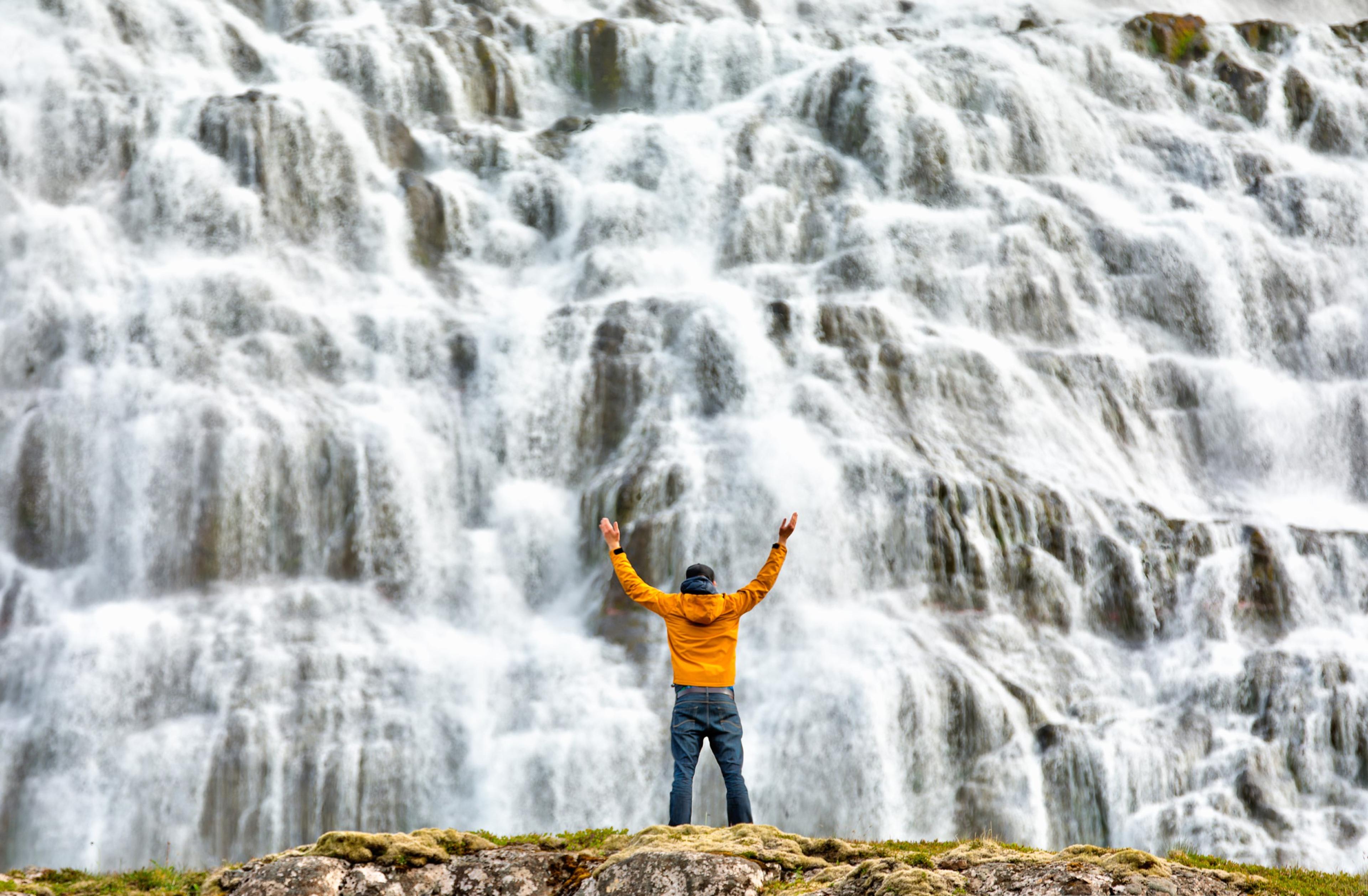 A person standing in front of a wide, white cascade with hands raised into the air