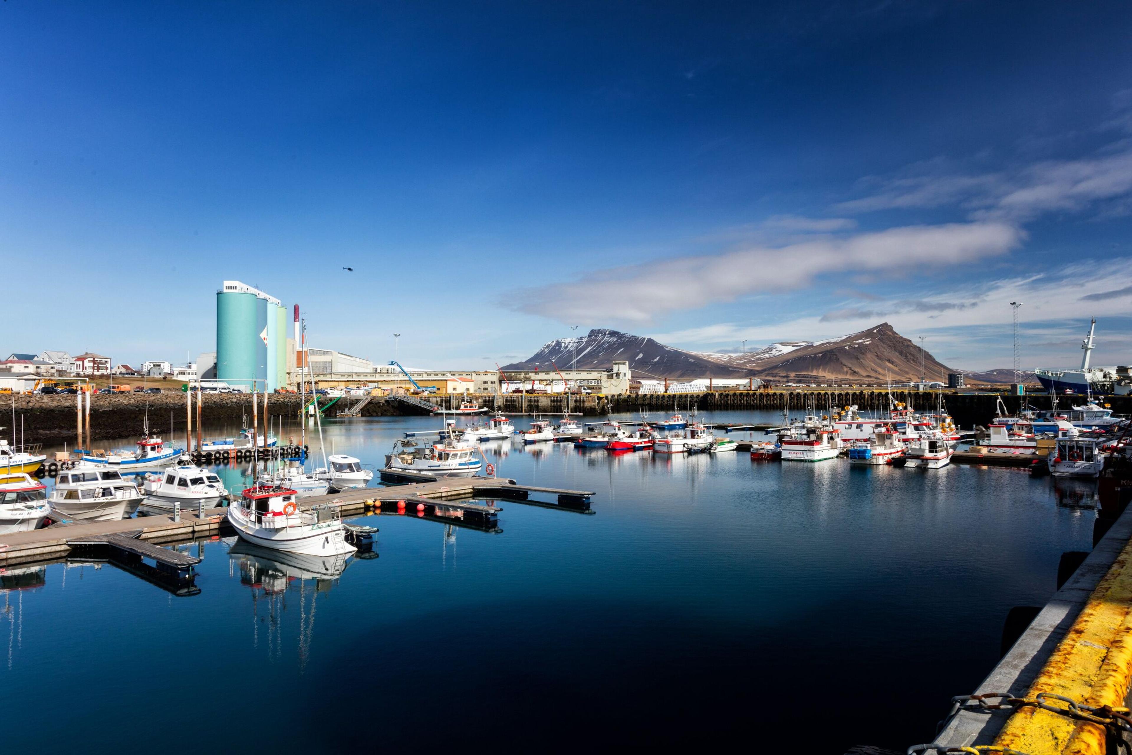 The harbor in Akranes, featuring docked fishing boats reflected in calm waters, with the town's buildings and distant snow-capped mountains under a clear blue sky.