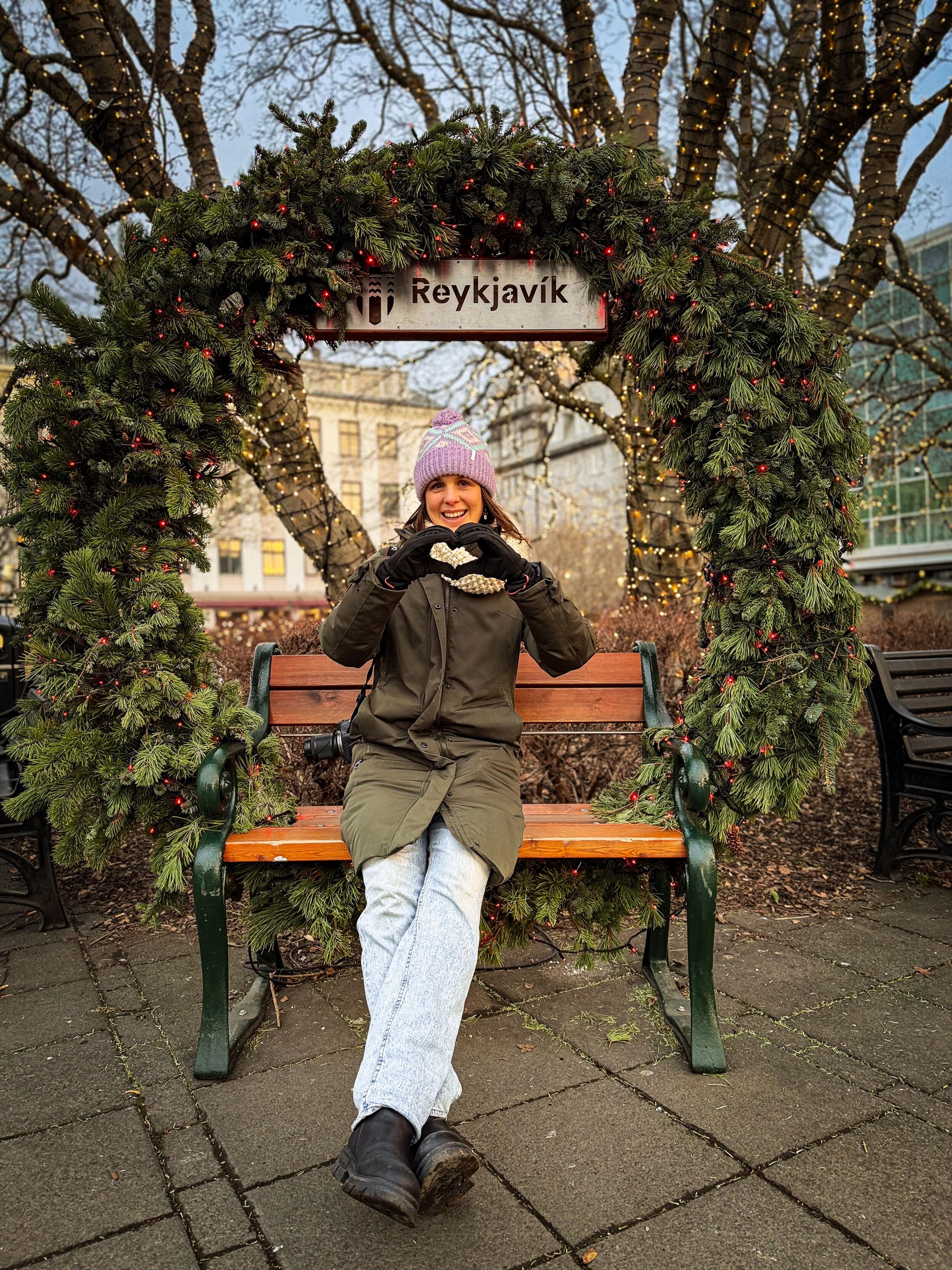 A woman in winter clothing sits on a bench, making a heart with her hands, under a festive arch with a "Reykjavík" sign.