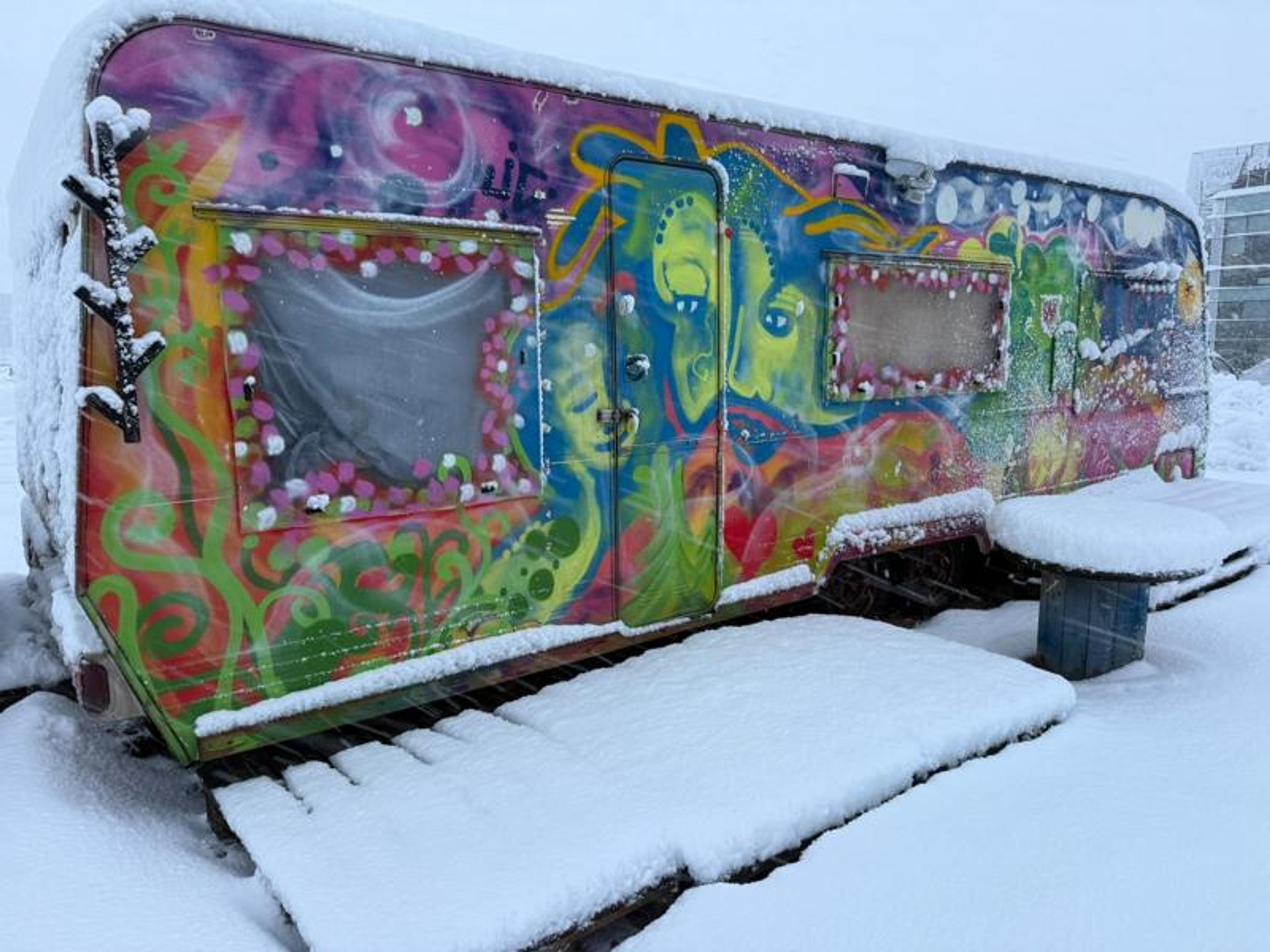 A colorful, graffiti-painted trailer covered in snow, with a snowy table and stool beside it.