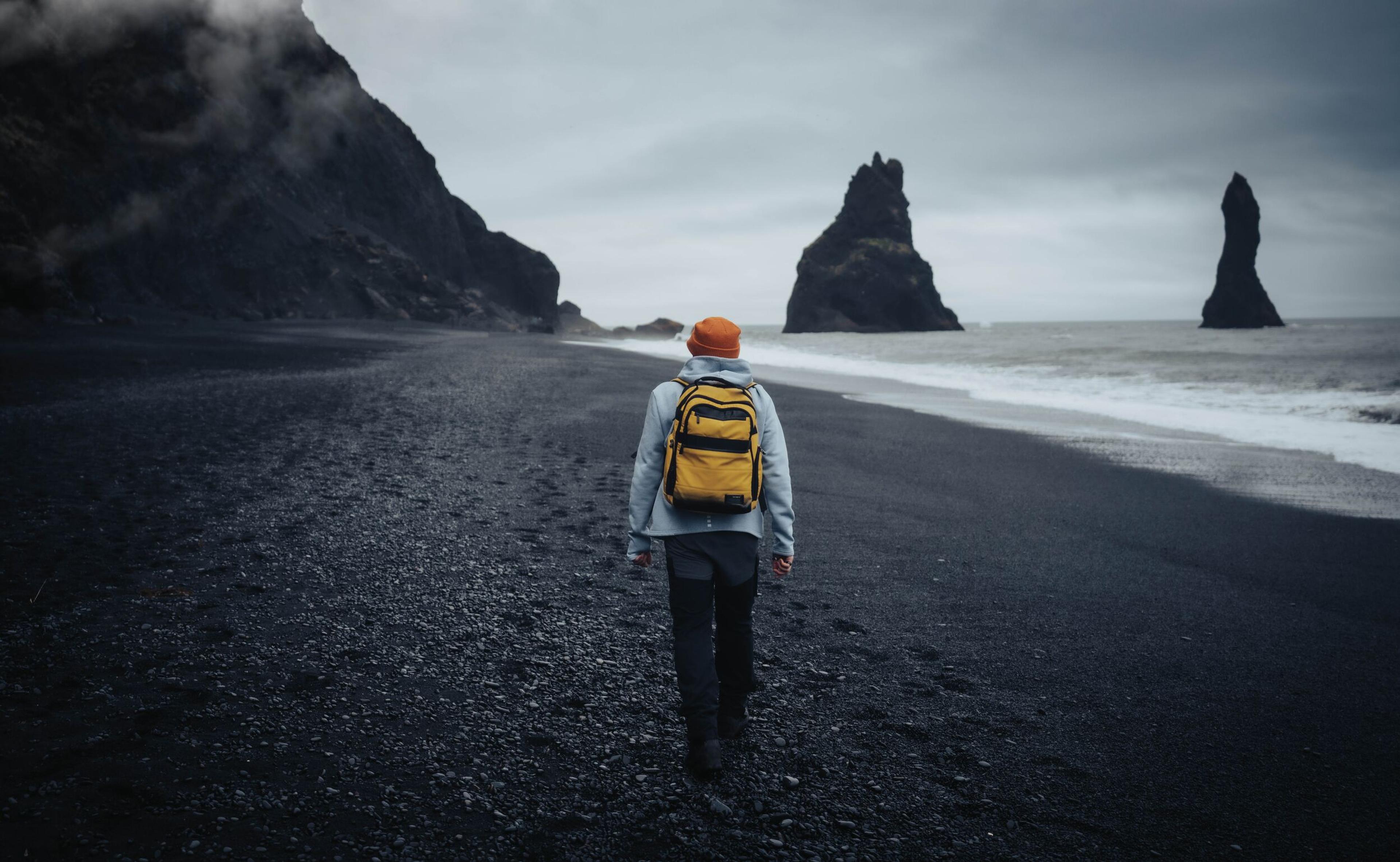 A person with a yellow backpack strolling on a black sand beach, heading towards the sea stacks in the distance.