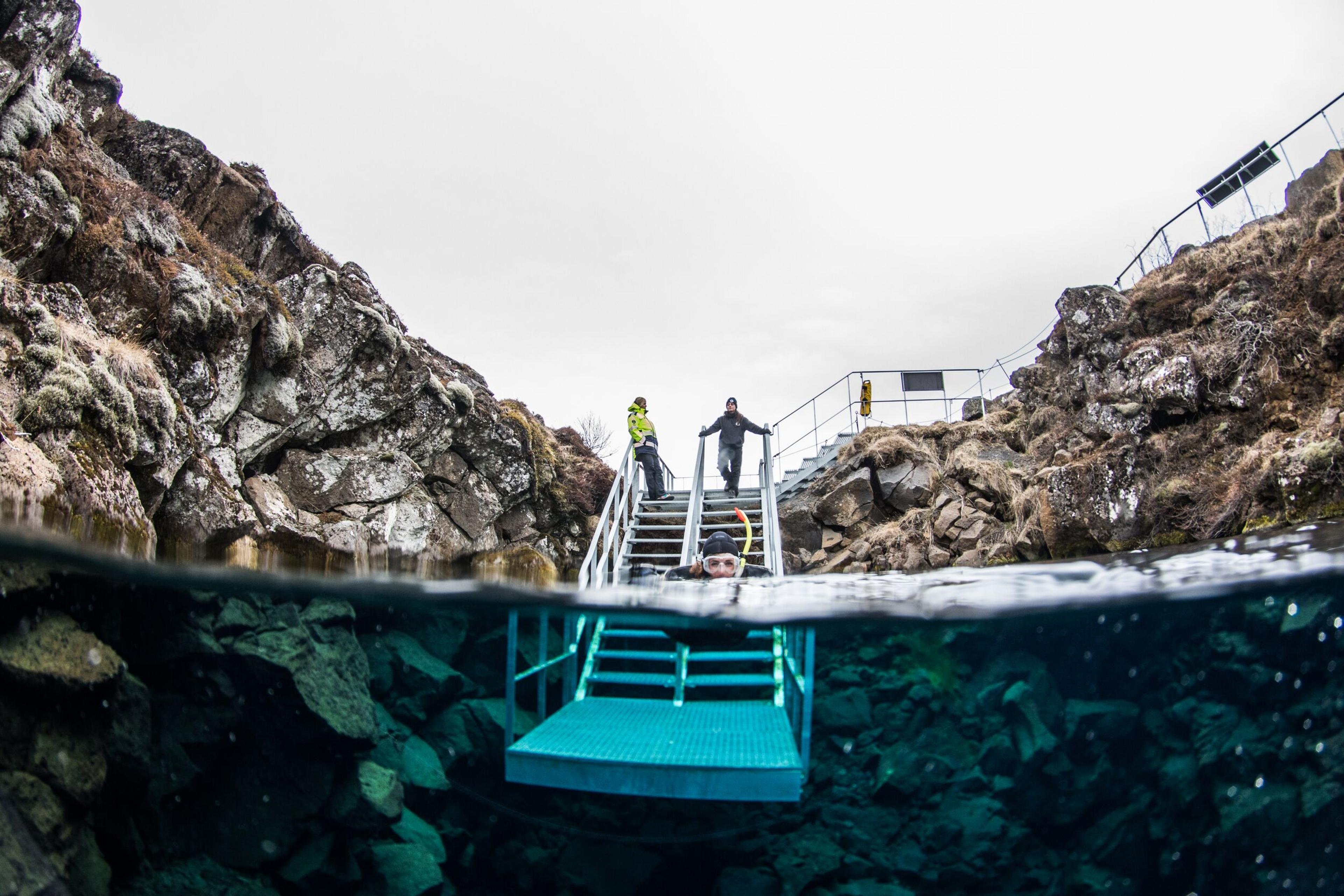 Half-submerged view of a snorkeler in clear water beneath a metal staircase, with rocky cliffs and onlookers above.