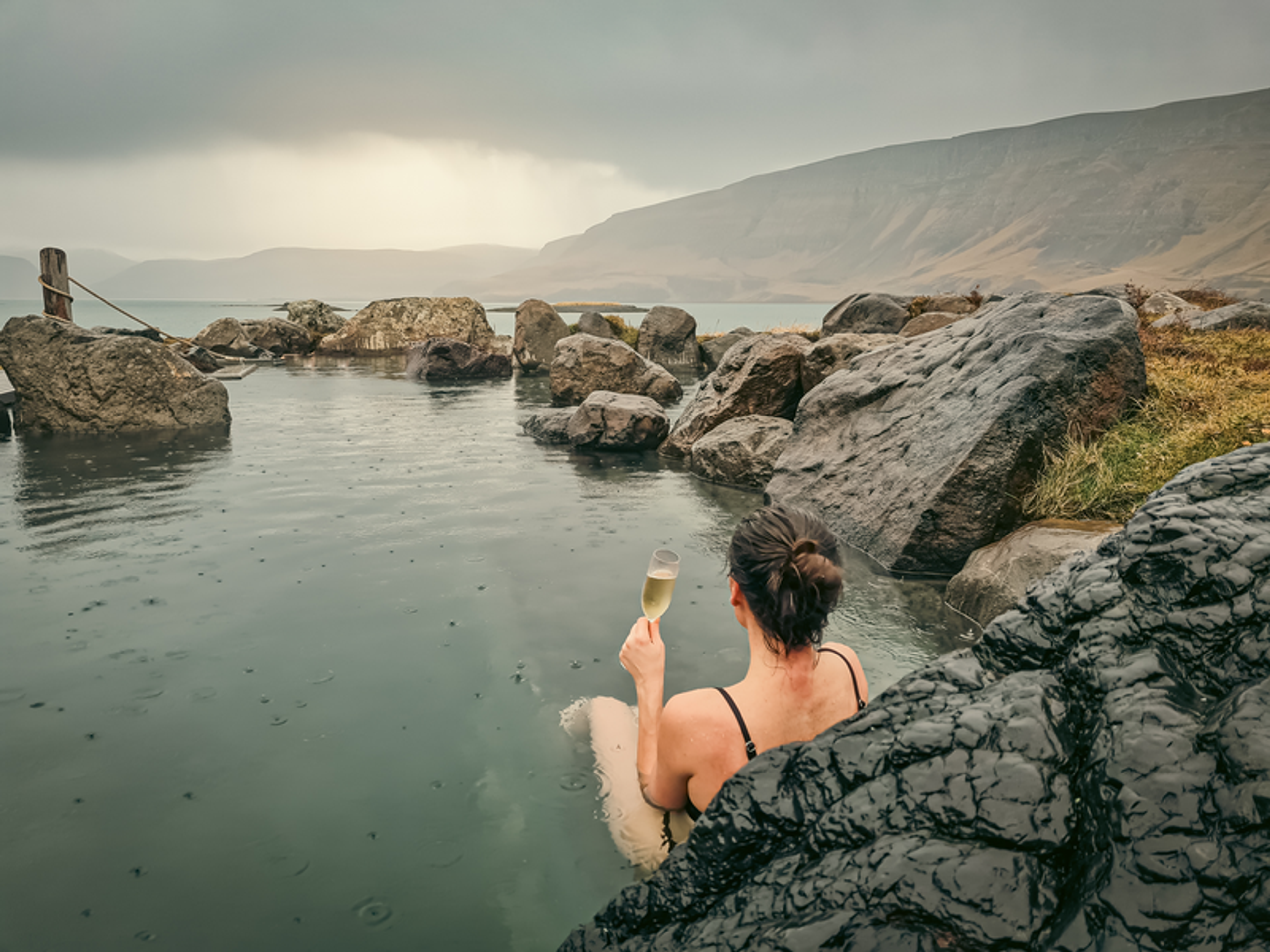Person relaxing in a geothermal pool surrounded by rocks, holding a beverage, with mountains in the background.