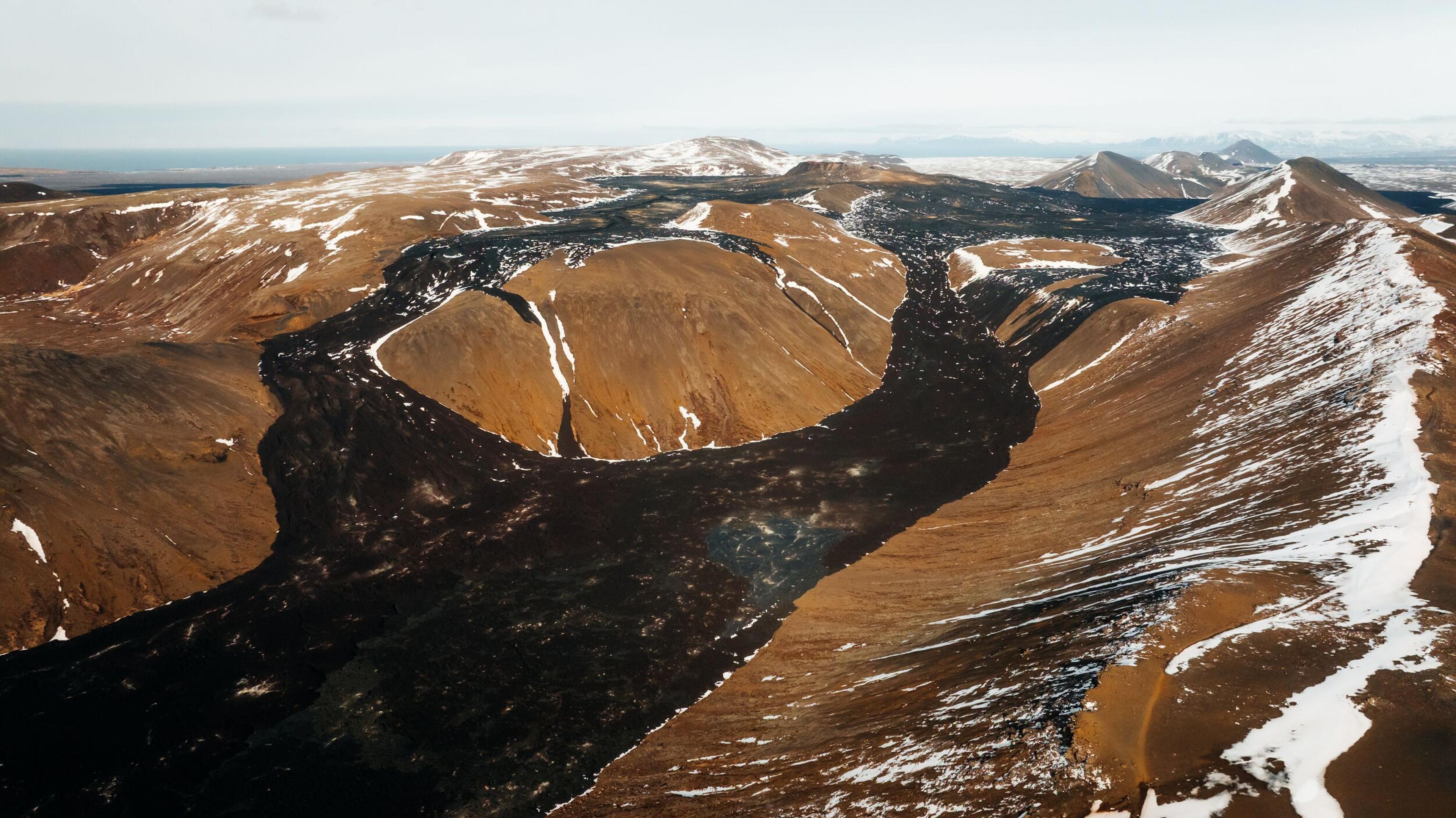 Aerial view of a dark lava flow snaking through snowy, reddish-brown volcanic hills.
