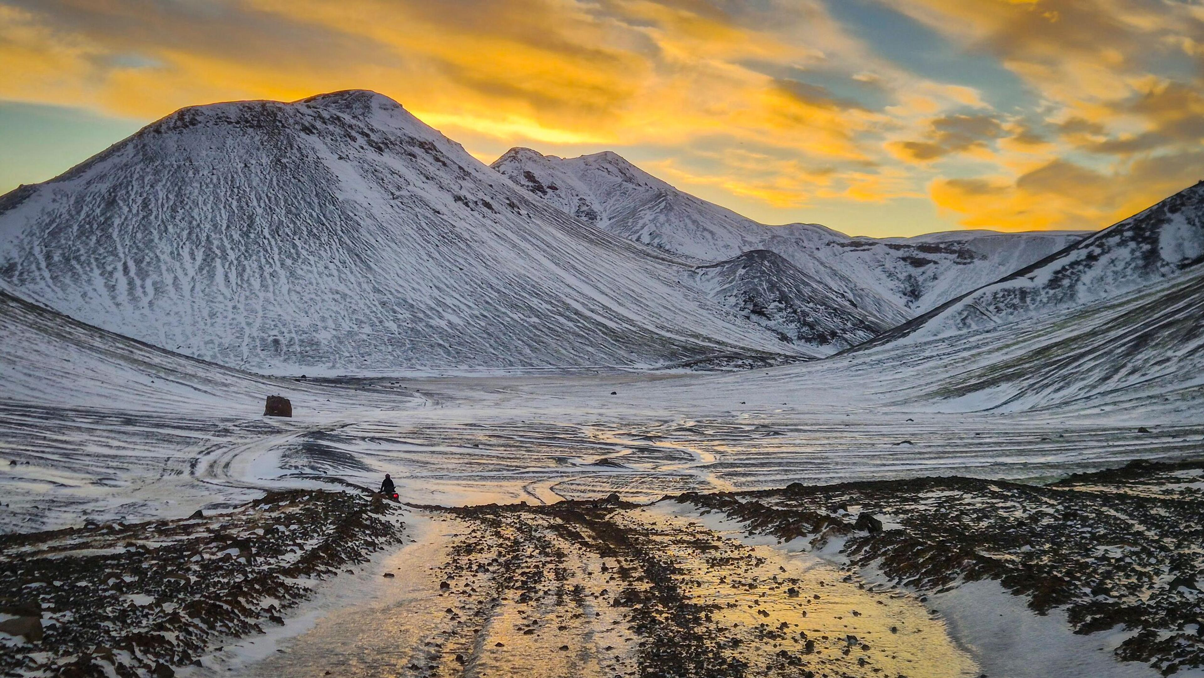 Snowy mountains surround a valley with a dirt road and patches of dark ground under a vibrant orange and yellow sky.