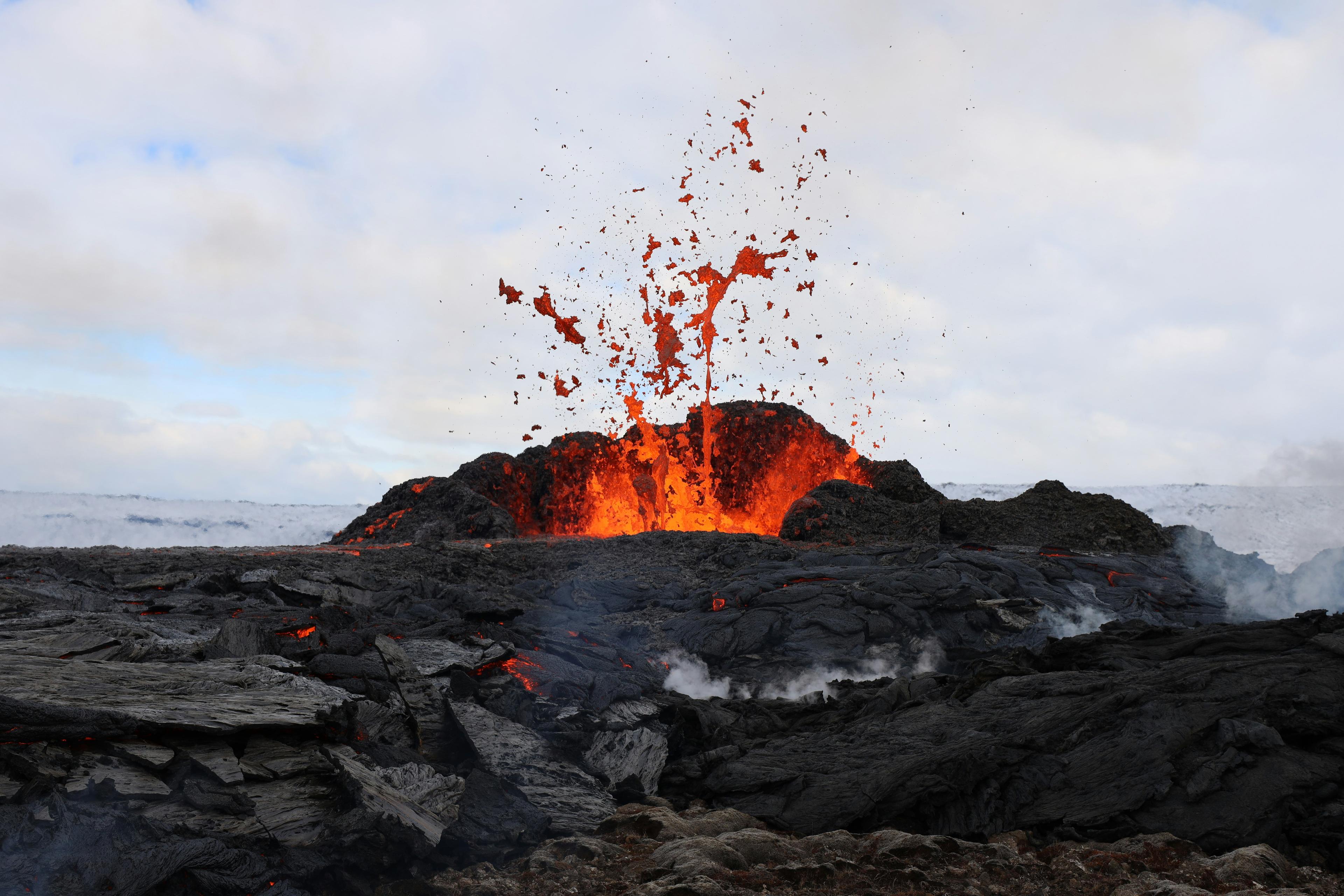 A volcanic eruption with bright orange lava spewing from a fissure, surrounded by dark, jagged rocks under a cloudy sky.