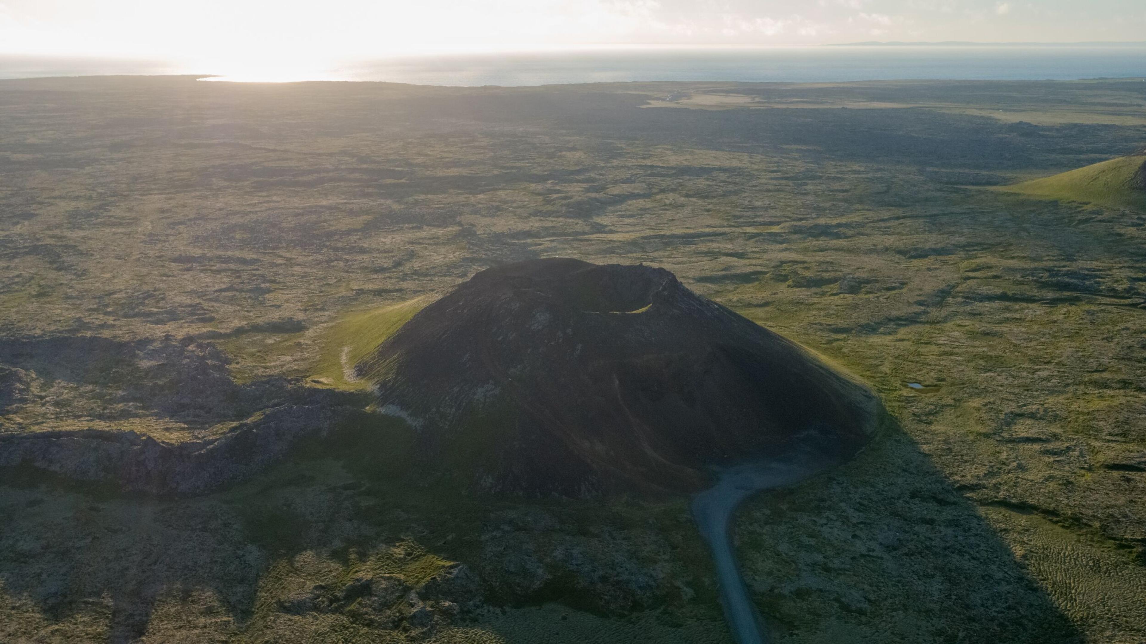 Aerial view of a dark volcanic cone with a crater in a green moss-covered landscape, with the ocean visible in the background.