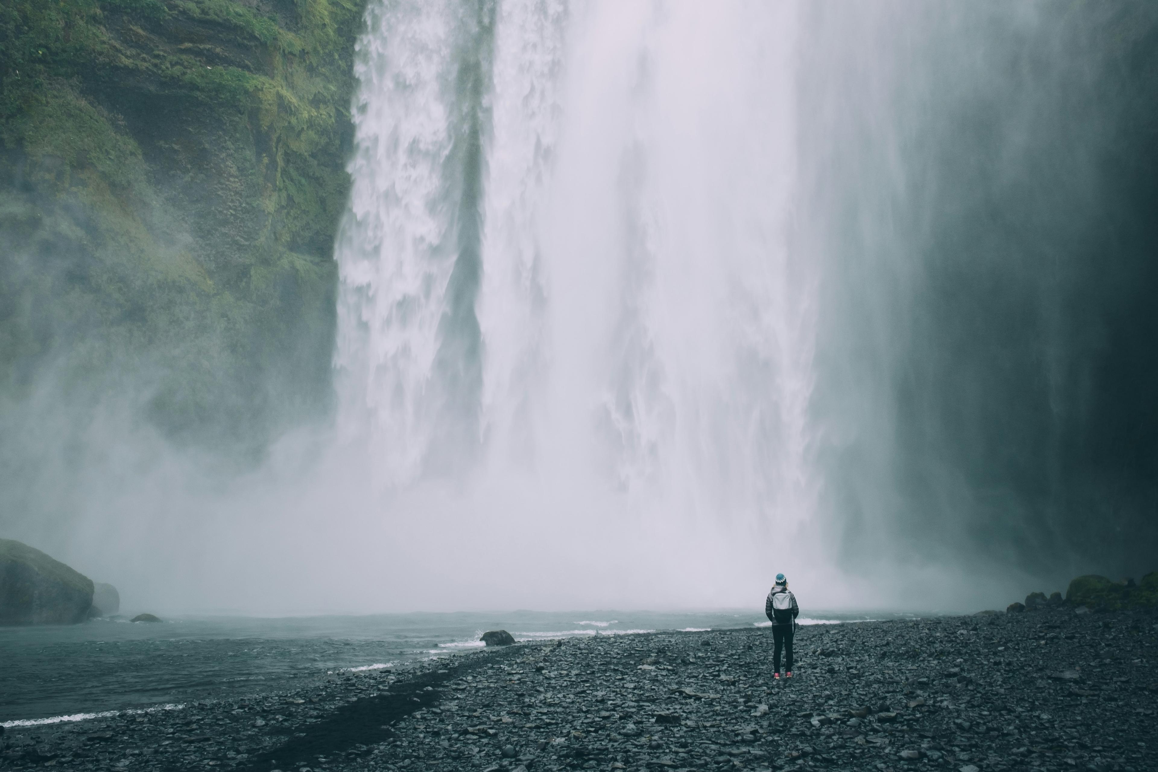 Person standing on rocky ground in front of a massive, misty waterfall in Iceland.