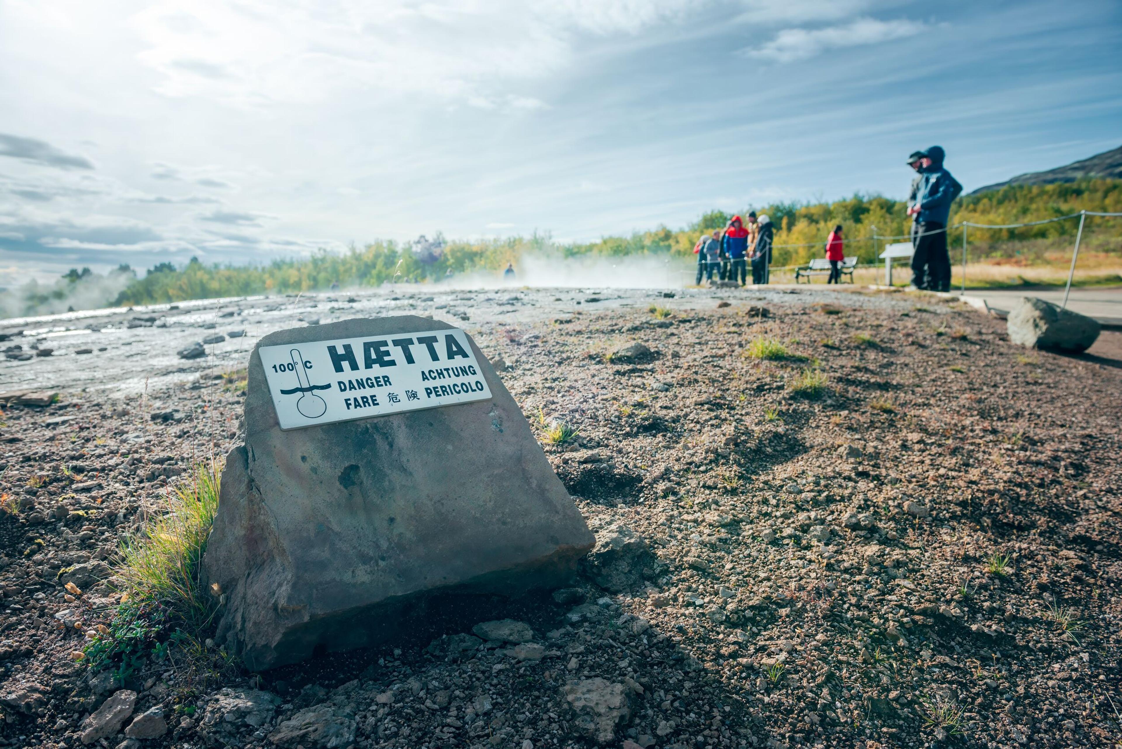 A close-up view of a geothermal warning sign in the foreground with unreadable text, with a blurred background of visitors walking on a path in the geothermal area of the Golden Circle, Iceland, under a bright sky