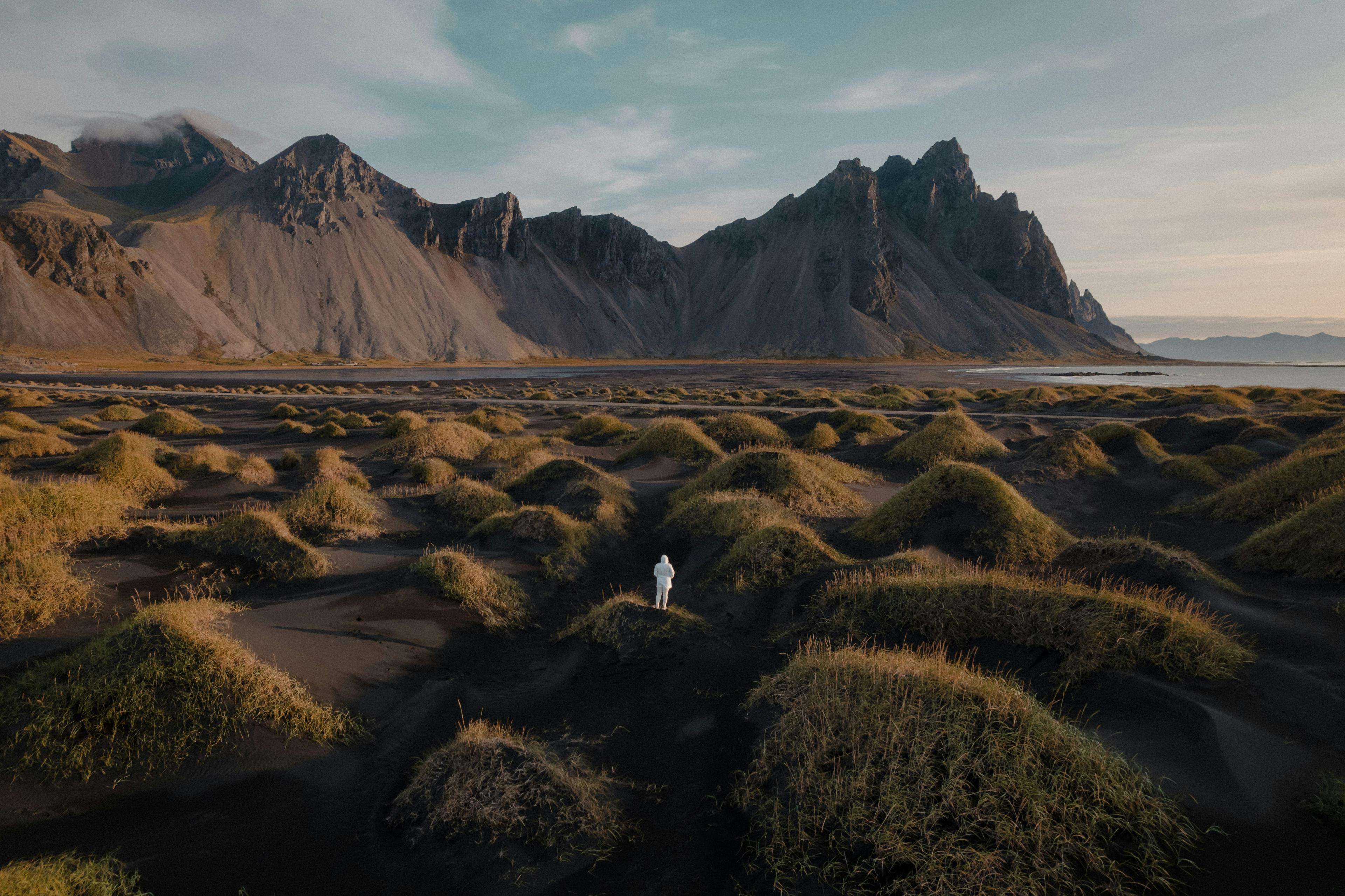 This image showcases the stunning Stokksnes peninsula in Iceland, featuring black sand dunes covered with tufts of green grass in the foreground. A solitary figure stands amidst the dramatic landscape, with sharp, jagged mountains towering in the background under a softly lit sky. 