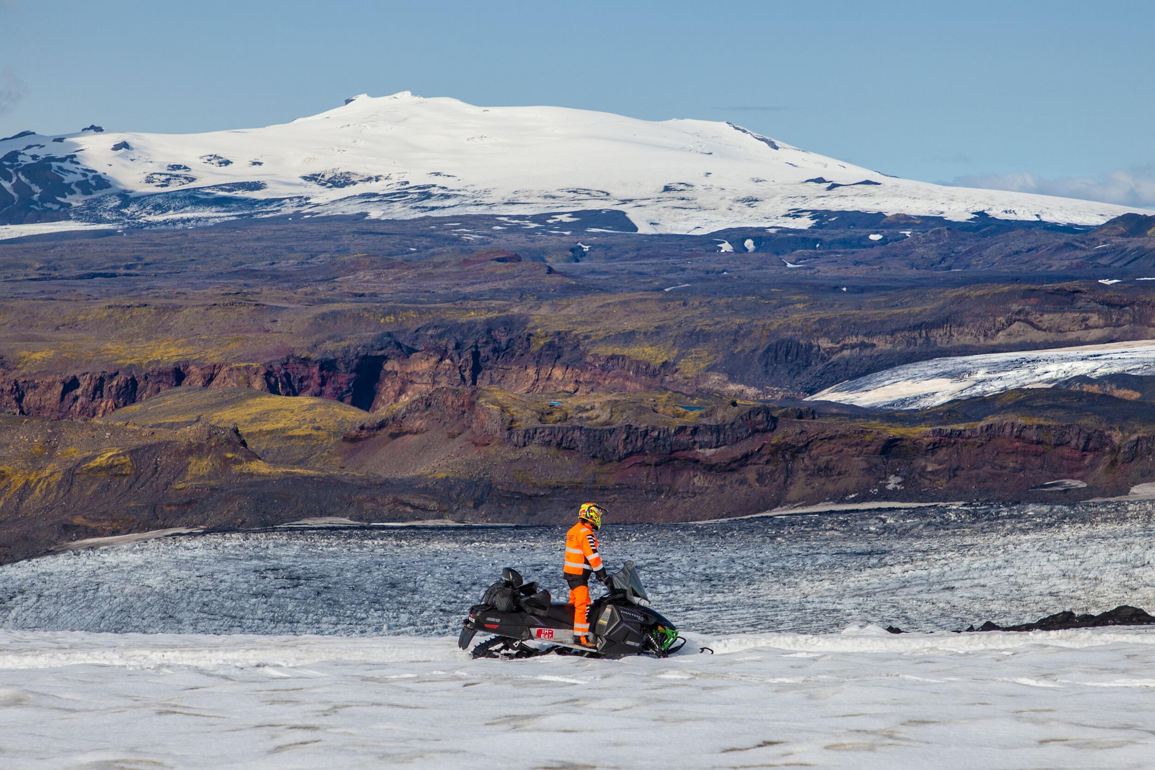 A person in high-visibility gear standing next to a snowmobile on a snowy expanse, with a backdrop of rugged cliffs and a snow-capped mountain under a clear blue sky.