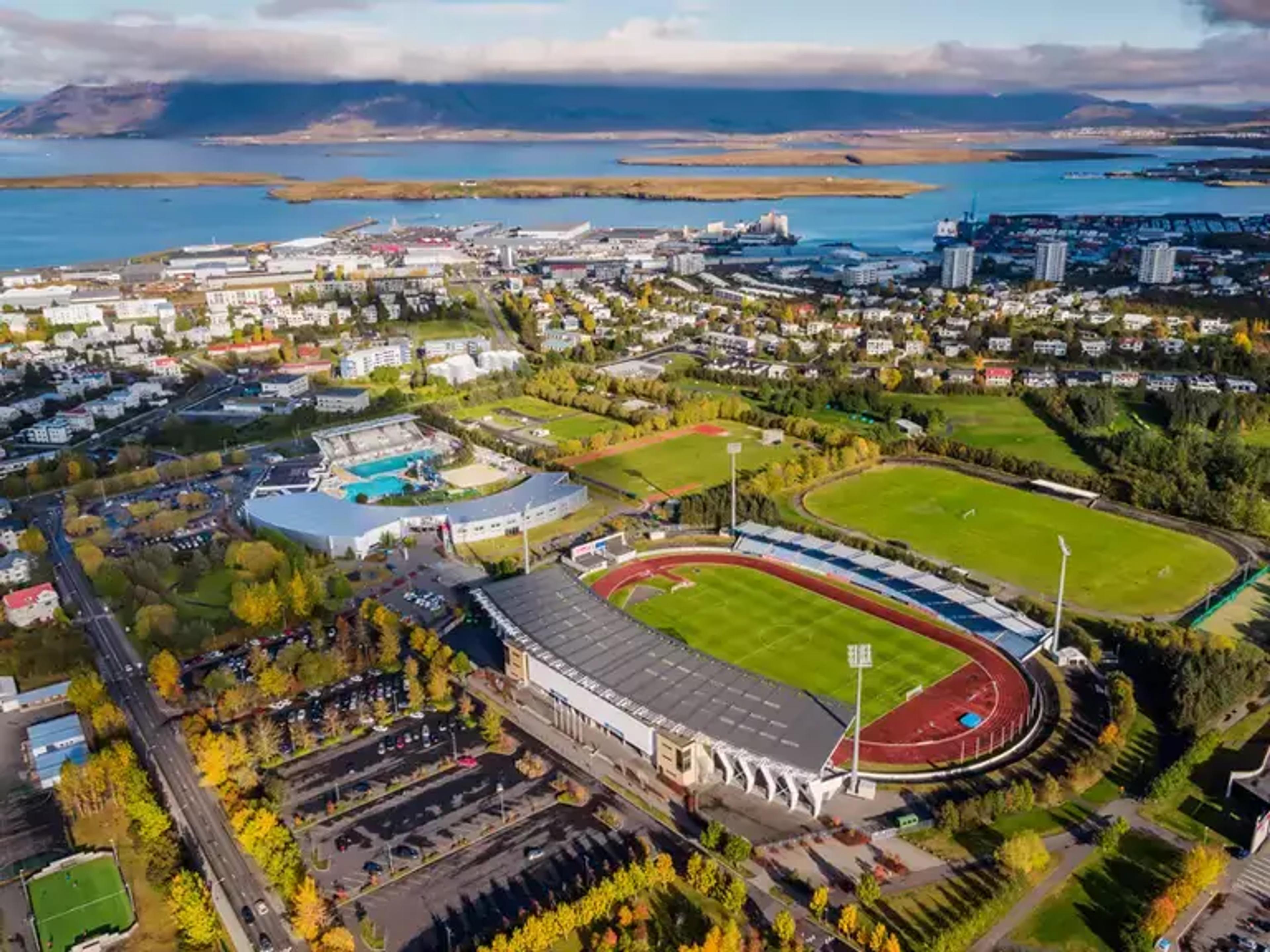 Aerial view of Laugardalsvöllur stadium surrounded by green spaces and Reykjavik cityscape with water in the background.