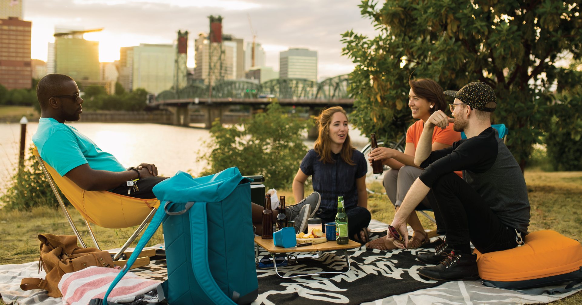Group of four people sitting in a park, laughing and talking, with a small wooden table set up in the middle with food, a speaker, a beer, and a small cylindrical silicone lantern