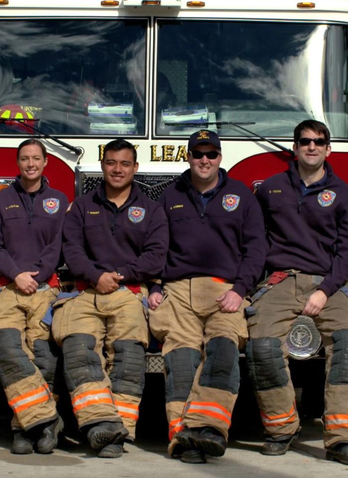 Group of four firefighters in uniform sitting in front of a fire truck, smiling for the photo.