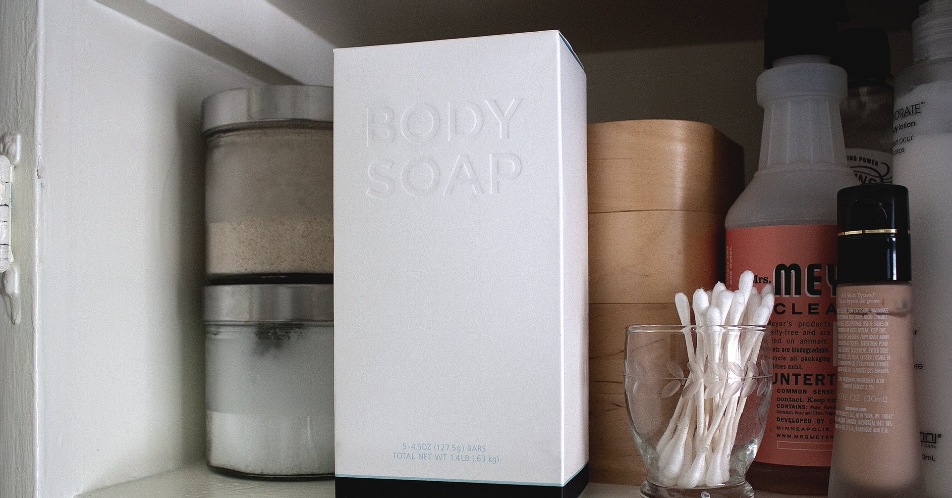 Plain white rectangular paper box that reads “Body Soap” in embossed lettering, sitting on a shelf in front of bathroom supplies and beauty products