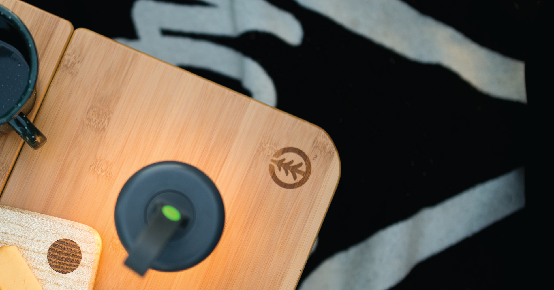 Small illuminated cylindrical LED lantern with a silicone holder on top, sitting on a wooden table that has a tree logo branded on the corner