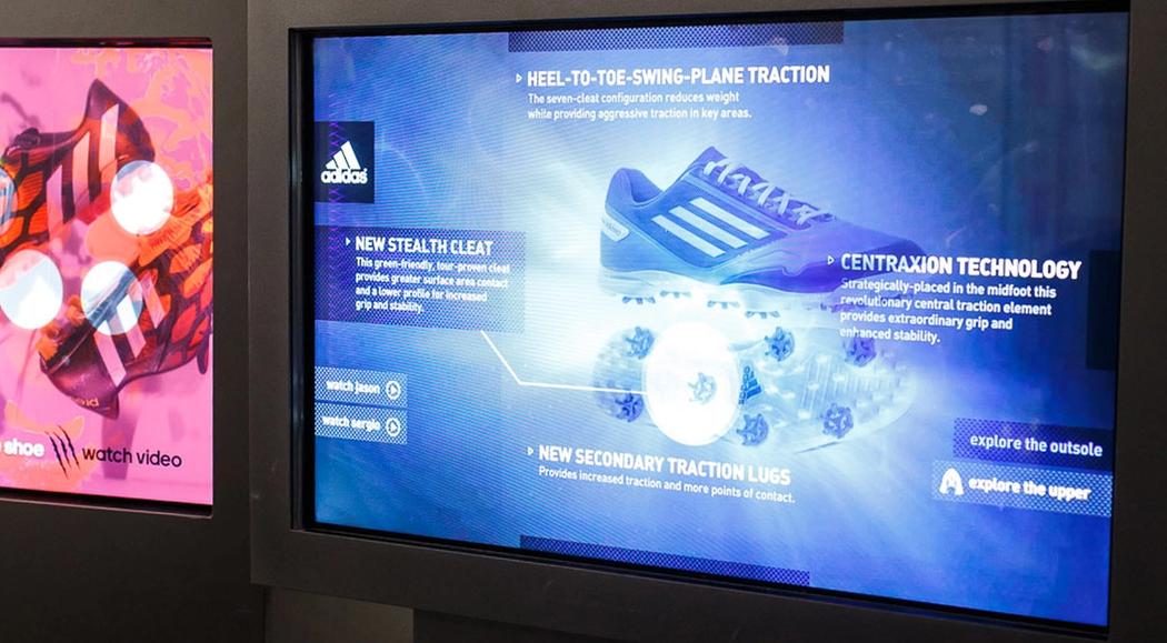 Blue touch screen showing technical and material details of two cleats that are suspended on string behind the screen