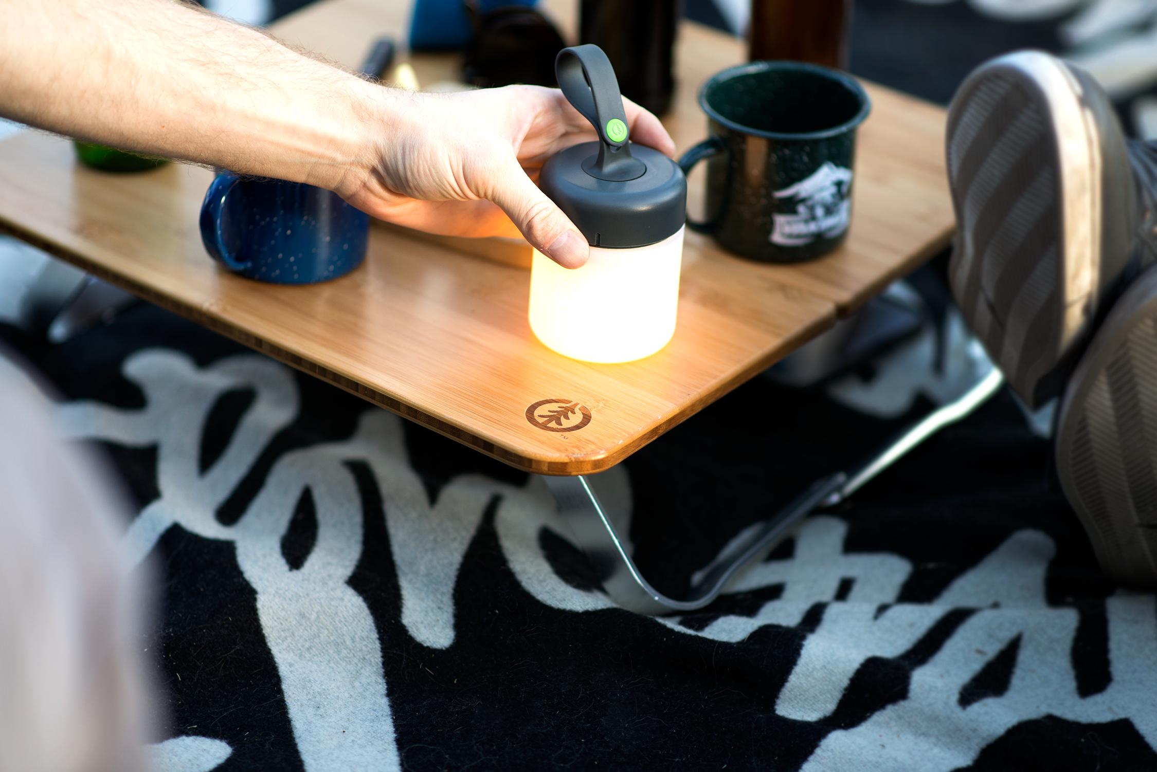 Person placing a small cylindrical silicone lantern next to two metal camping mugs on a small wooden table that has angled metal supports on two sides