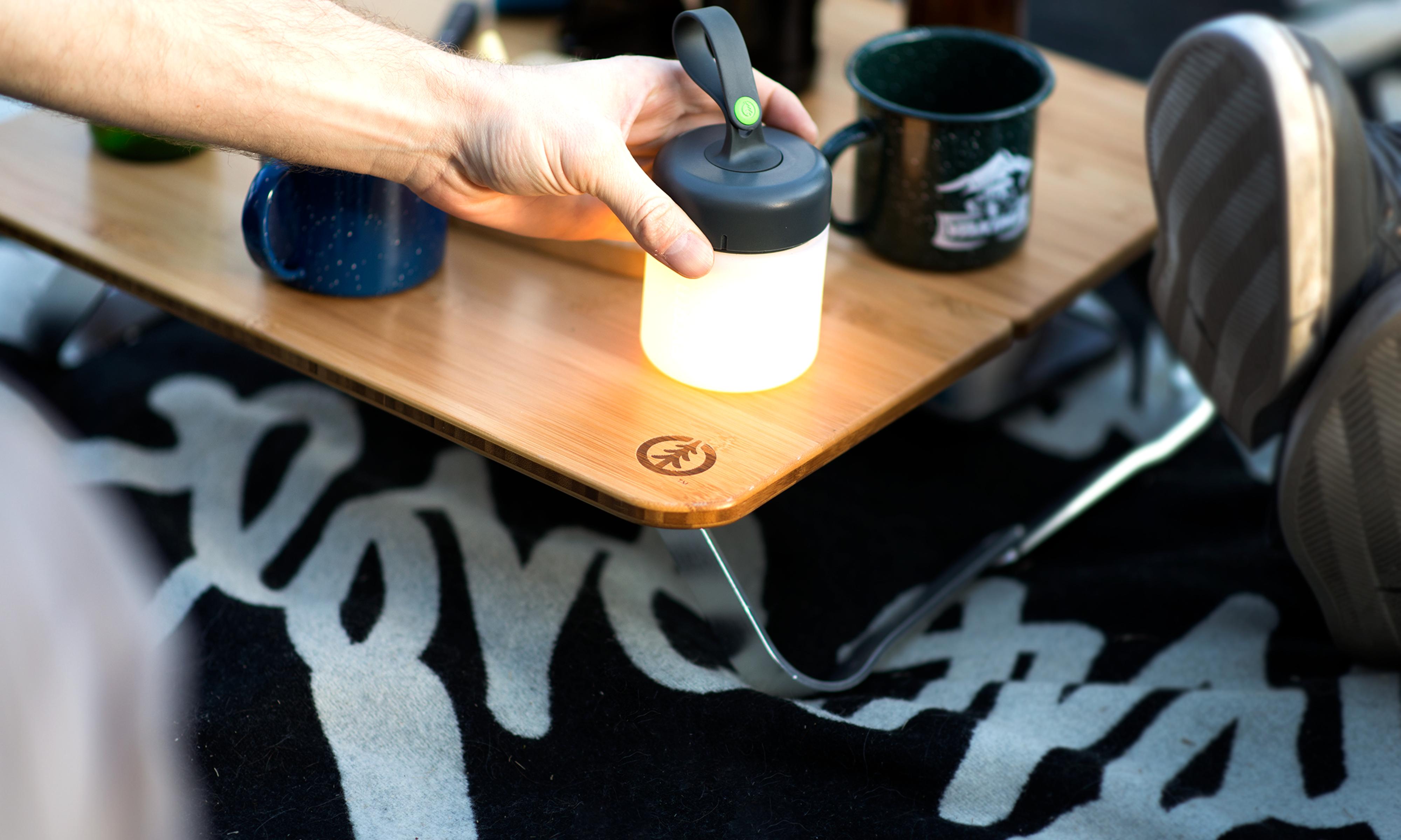 Person placing a small cylindrical silicone lantern next to two metal camping mugs on a small wooden table that has angled metal supports on two sides