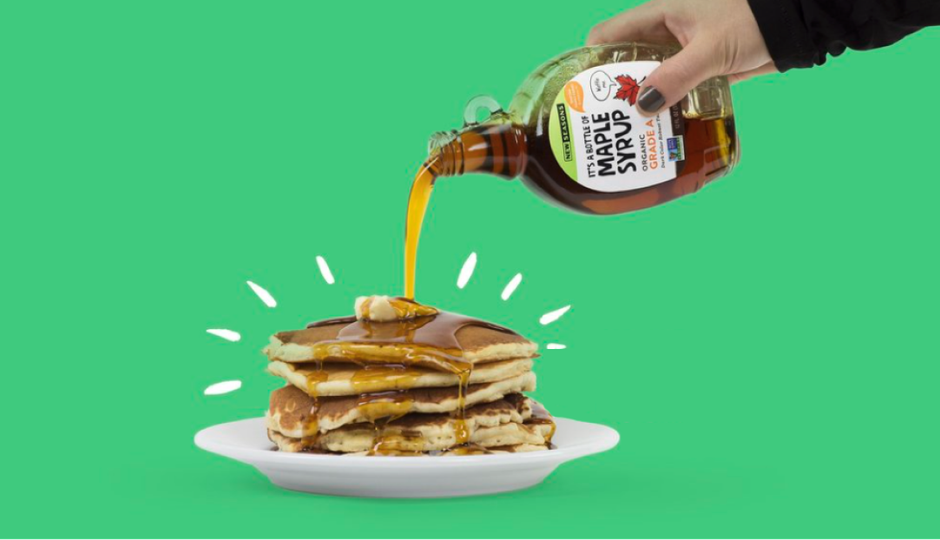 Person pouring maple syrup from a small glass container onto a stack of pancakes on a white plate, all in front of a green background